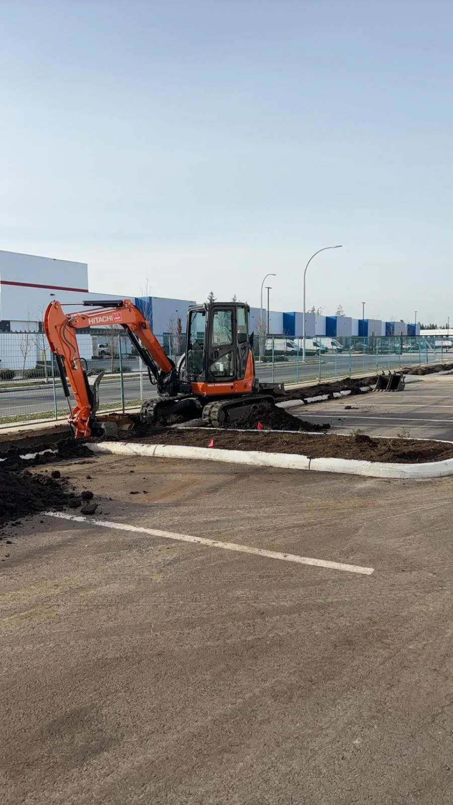 An orange excavator digging soil on a construction site in a parking lot with industrial buildings in the background.