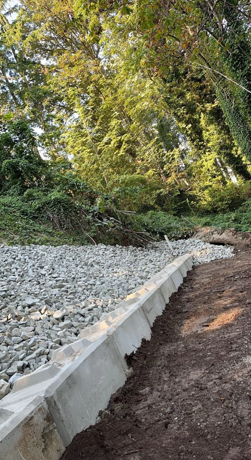 Concrete retaining wall with gravel and dirt path in wooded area with trees and foliage.