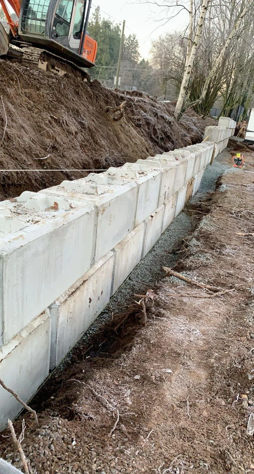 Excavator near concrete block retaining wall under construction with dirt and gravel in a wooded area.