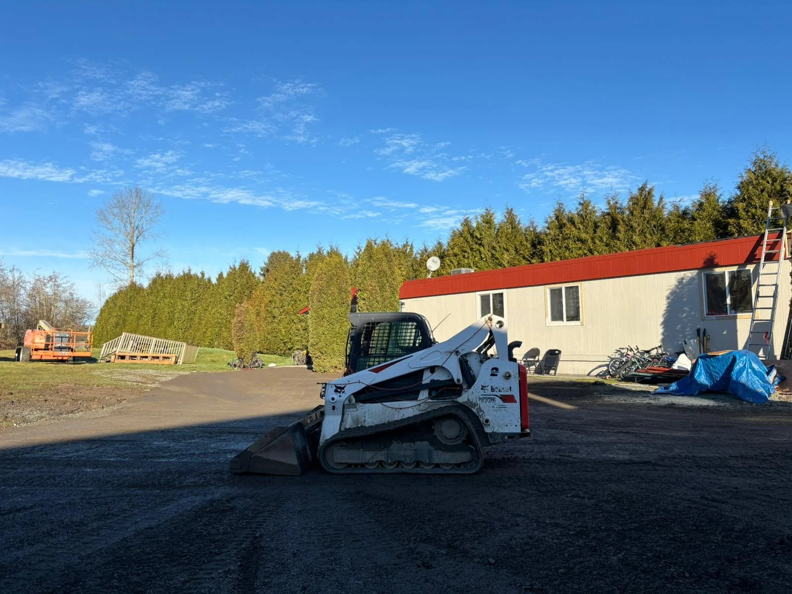 Outdoor scene with a compact track loader in front of a mobile office building, surrounded by trees and a ladder leaning against the structure.