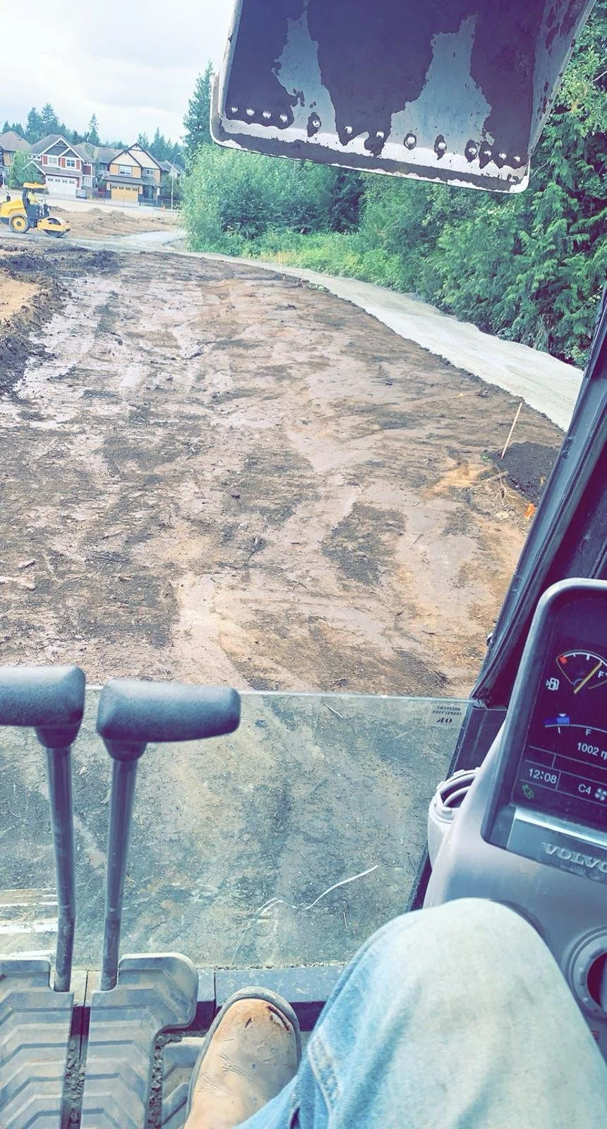 View from inside a construction vehicle showing muddy ground and a construction site with residential houses and trees in the background.