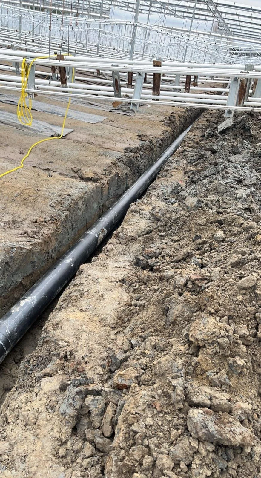 A greenhouse under construction with a trench dug in the ground, containing a black pipe and dirt piled alongside. Metal and plastic structural elements are visible, along with some yellow cables.
