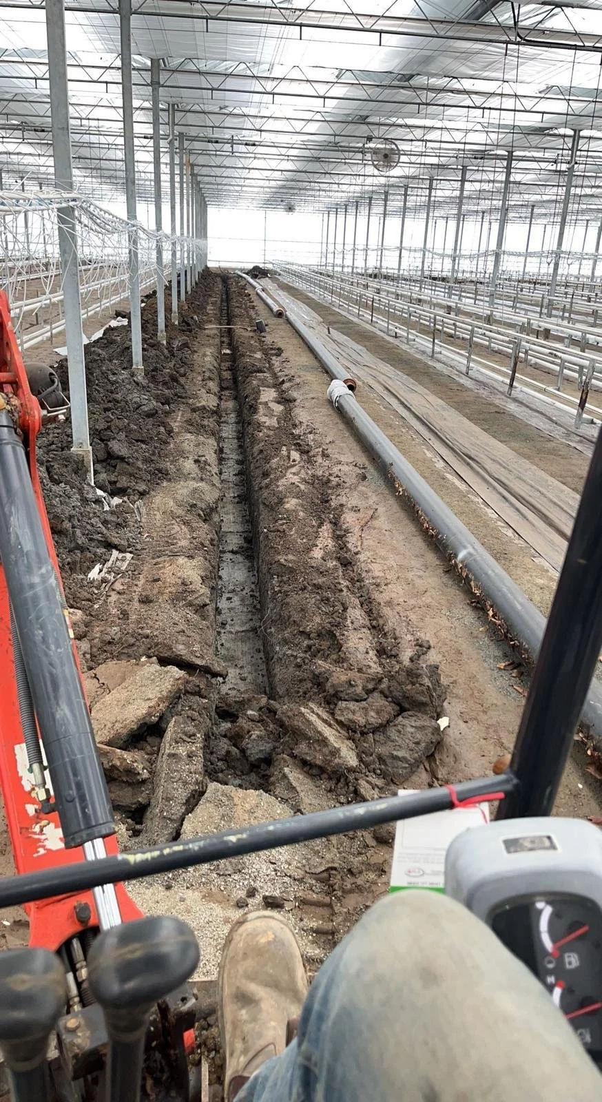 Interior of a greenhouse under construction, featuring a long trench with exposed soil and visible pipes. A red excavator is partially visible, likely being operated inside the greenhouse.