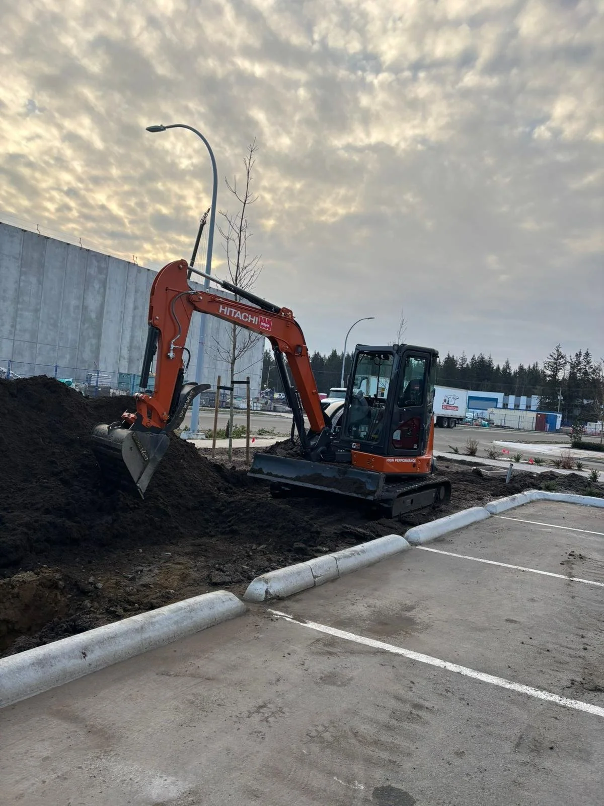 Small orange excavator digging near a parking lot with overcast sky