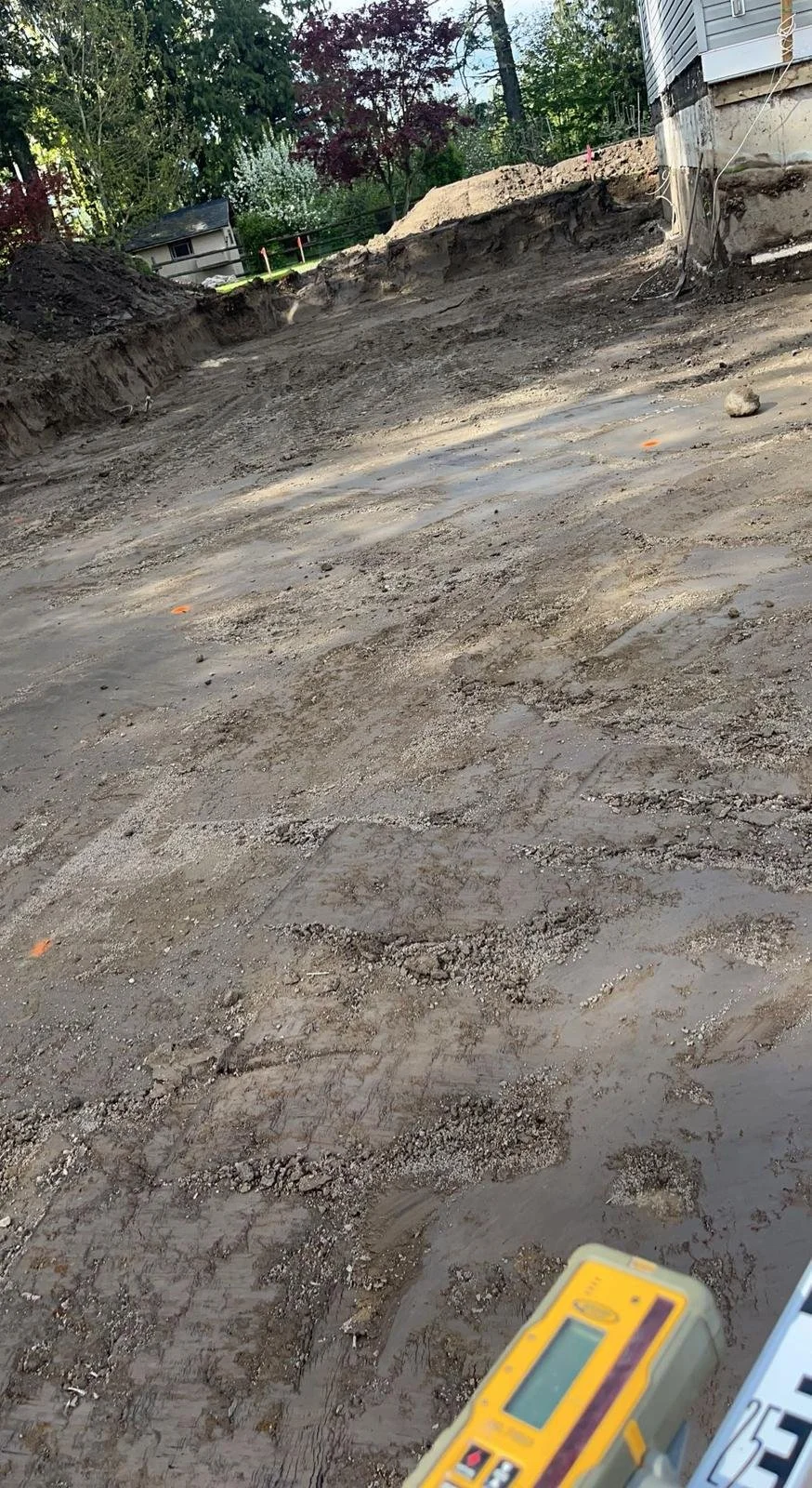 A construction site with a cleared dirt area, a house foundation, and surveying equipment in the foreground.