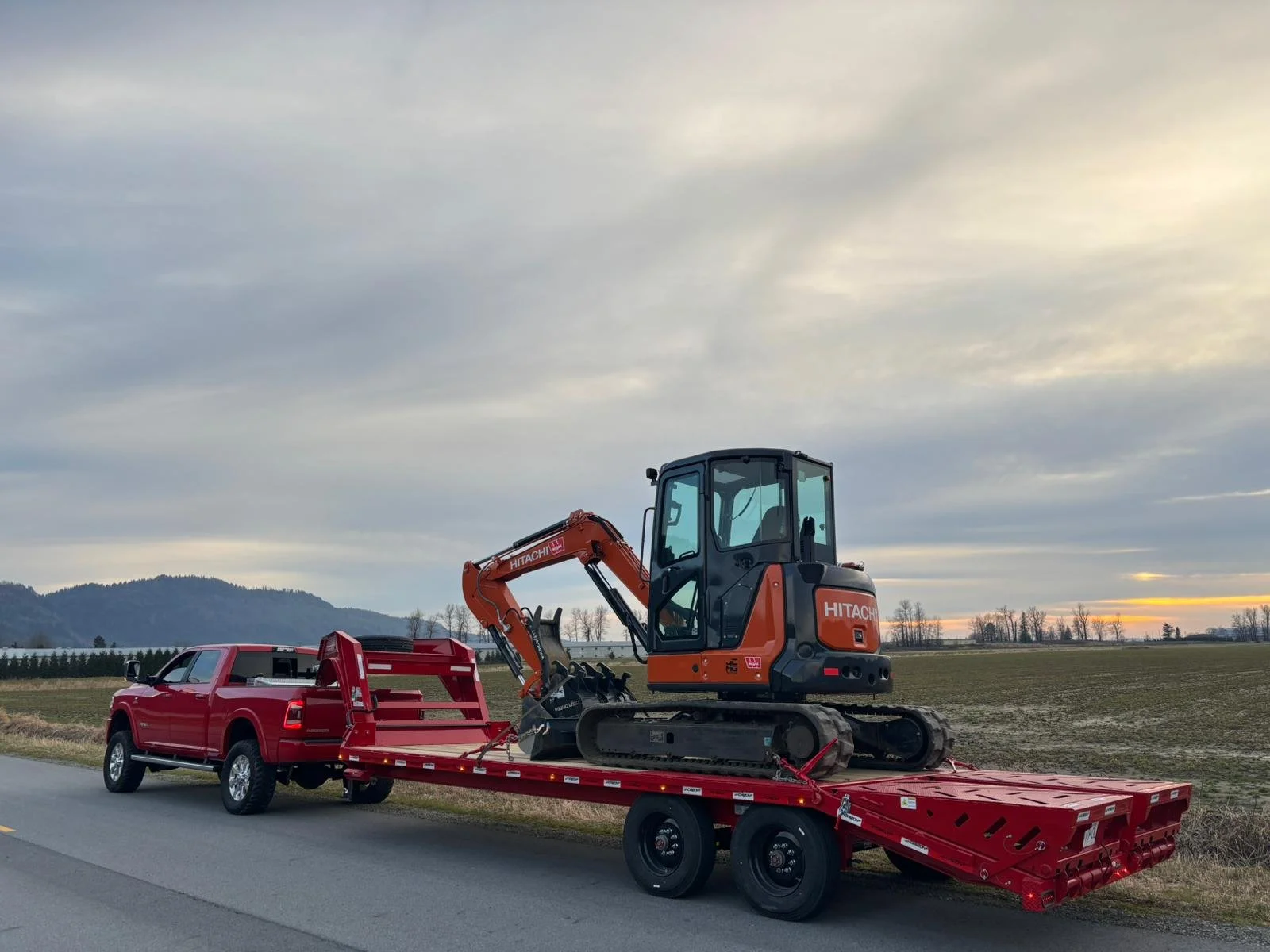 A red pickup truck towing a red flatbed trailer with an orange mini excavator on it, parked on a rural road with fields and hills in the background.