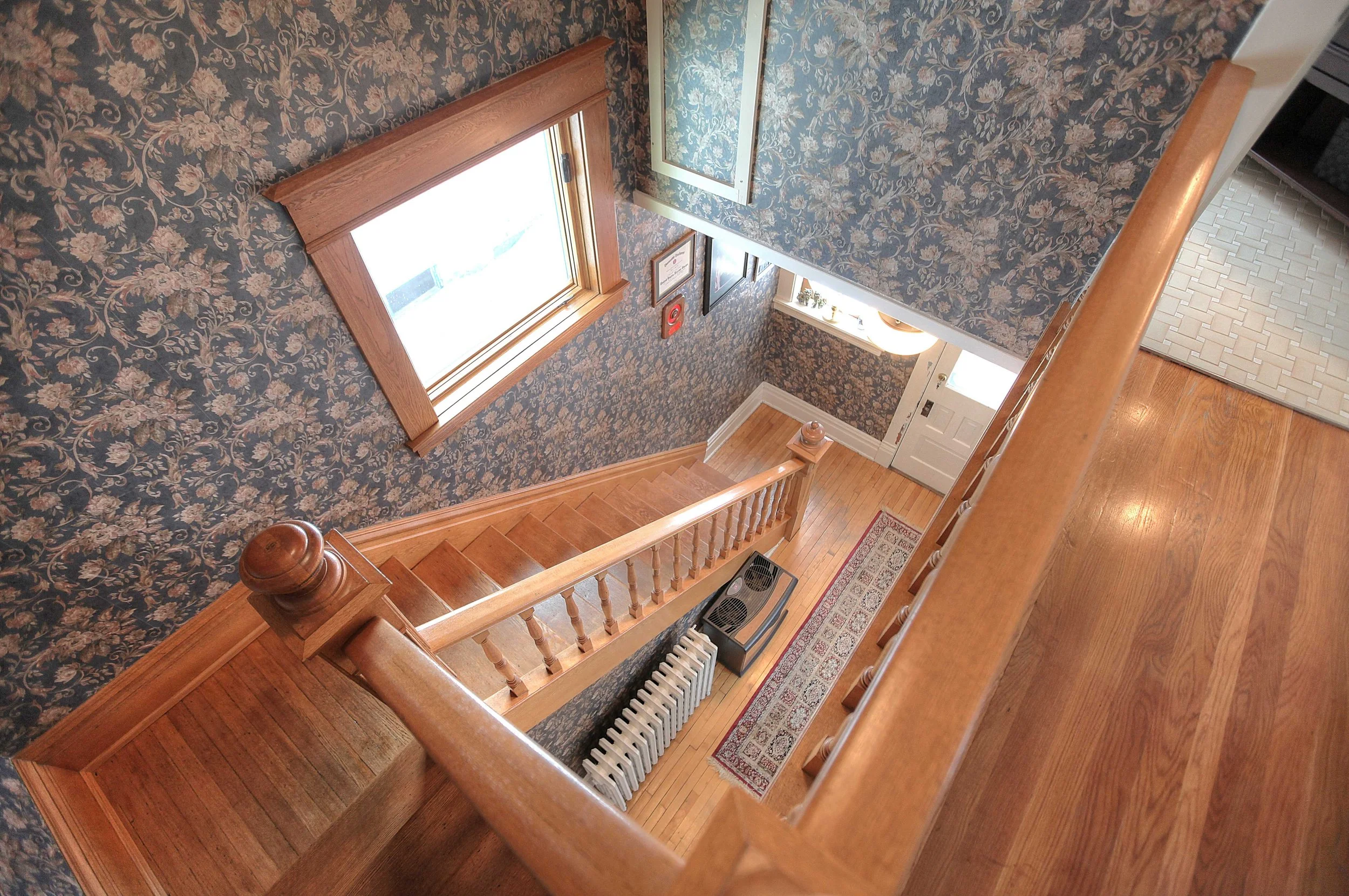 Wooden staircase in a house with floral wallpaper, leading to a hallway with a patterned rug, radiator, and a window with wooden trim.