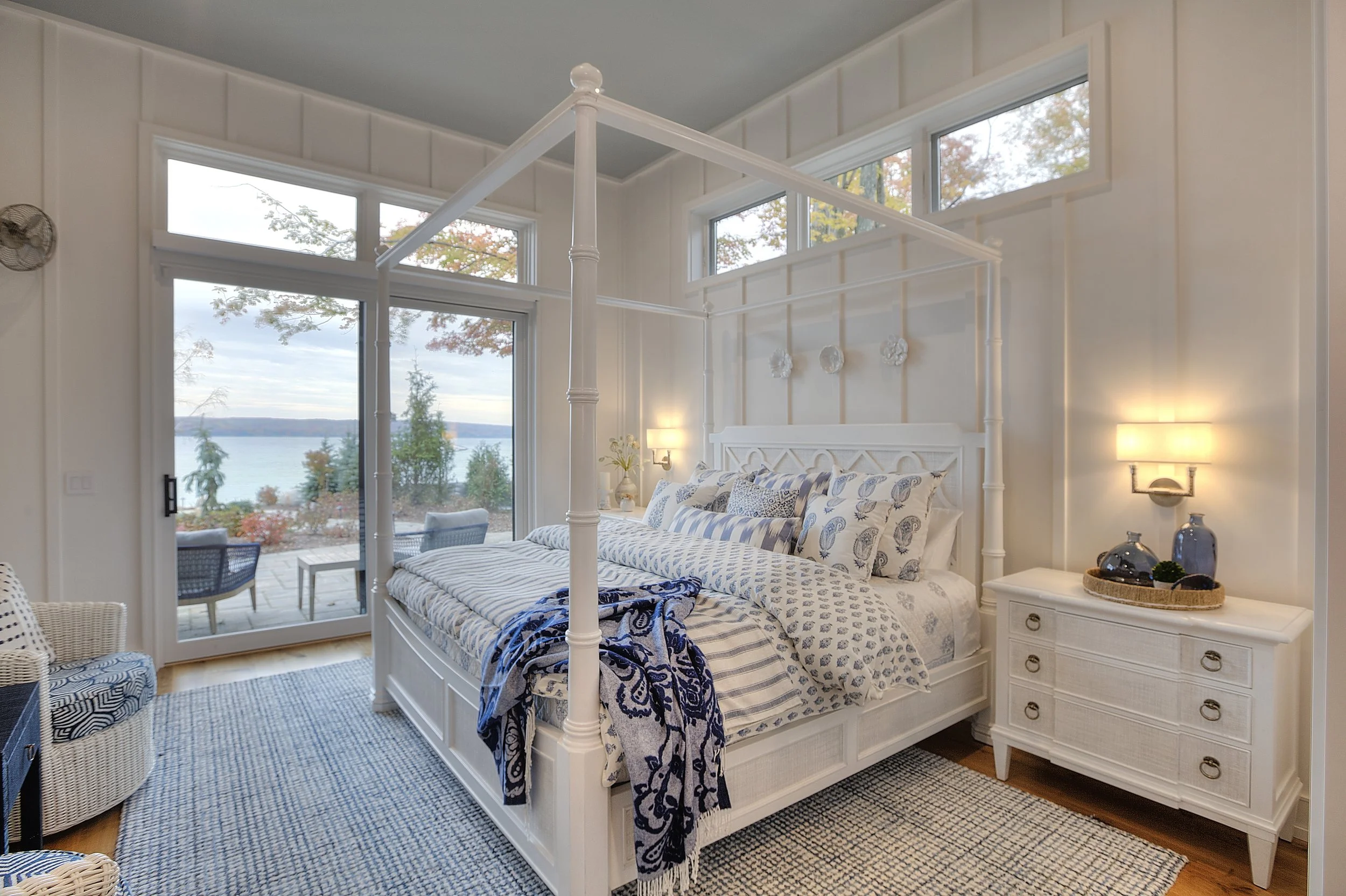 Bedroom with a four-poster bed, blue and white bedding, nightstand with lamp, and large windows with a lake view.