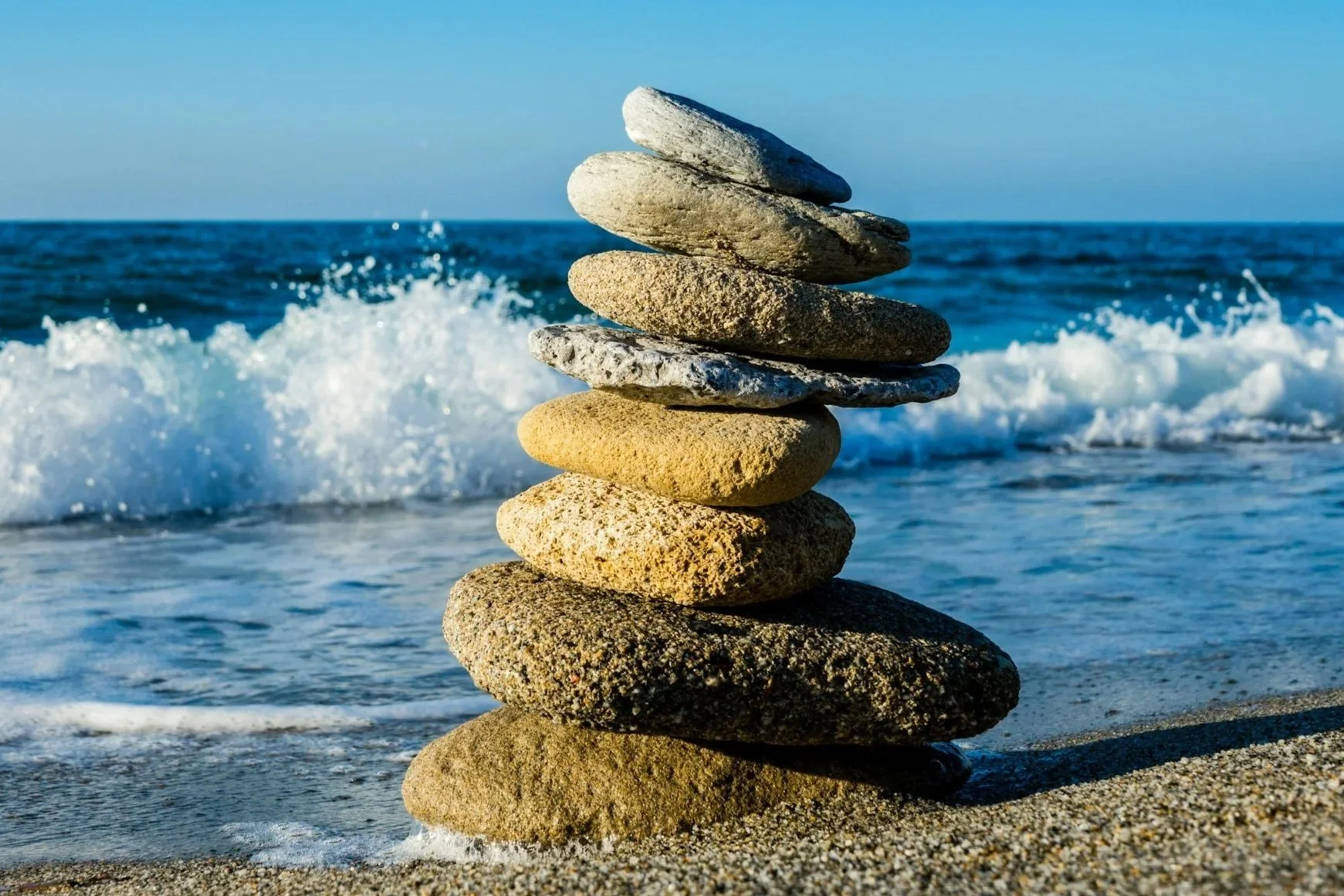 Stack of smooth stones on the sandy beach with ocean waves and blue sky in the background.