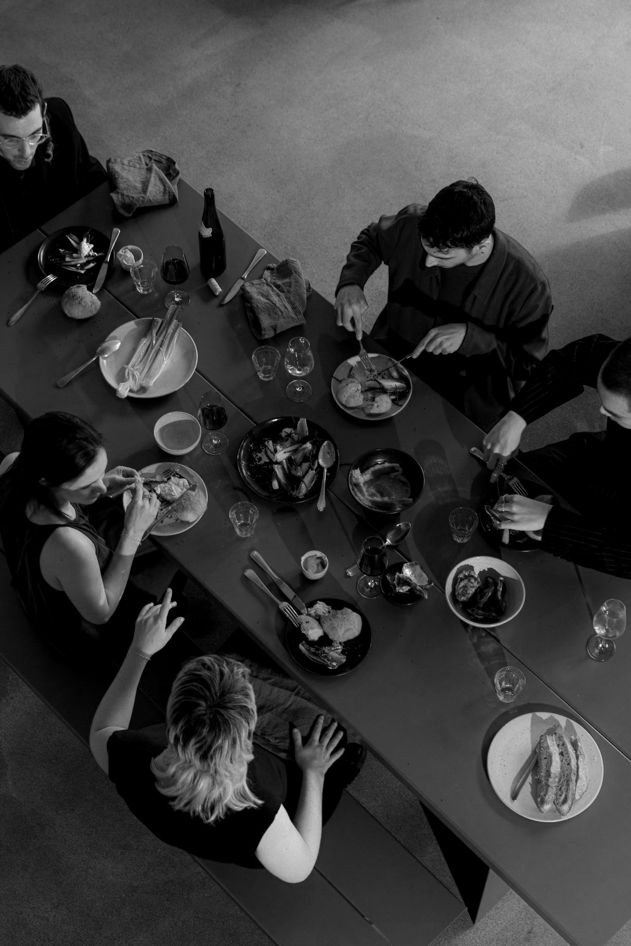 A black and white top-down view of a group of six people sitting around a long dining table, eating and drinking, with dishes, glasses, and utensils scattered across the table.