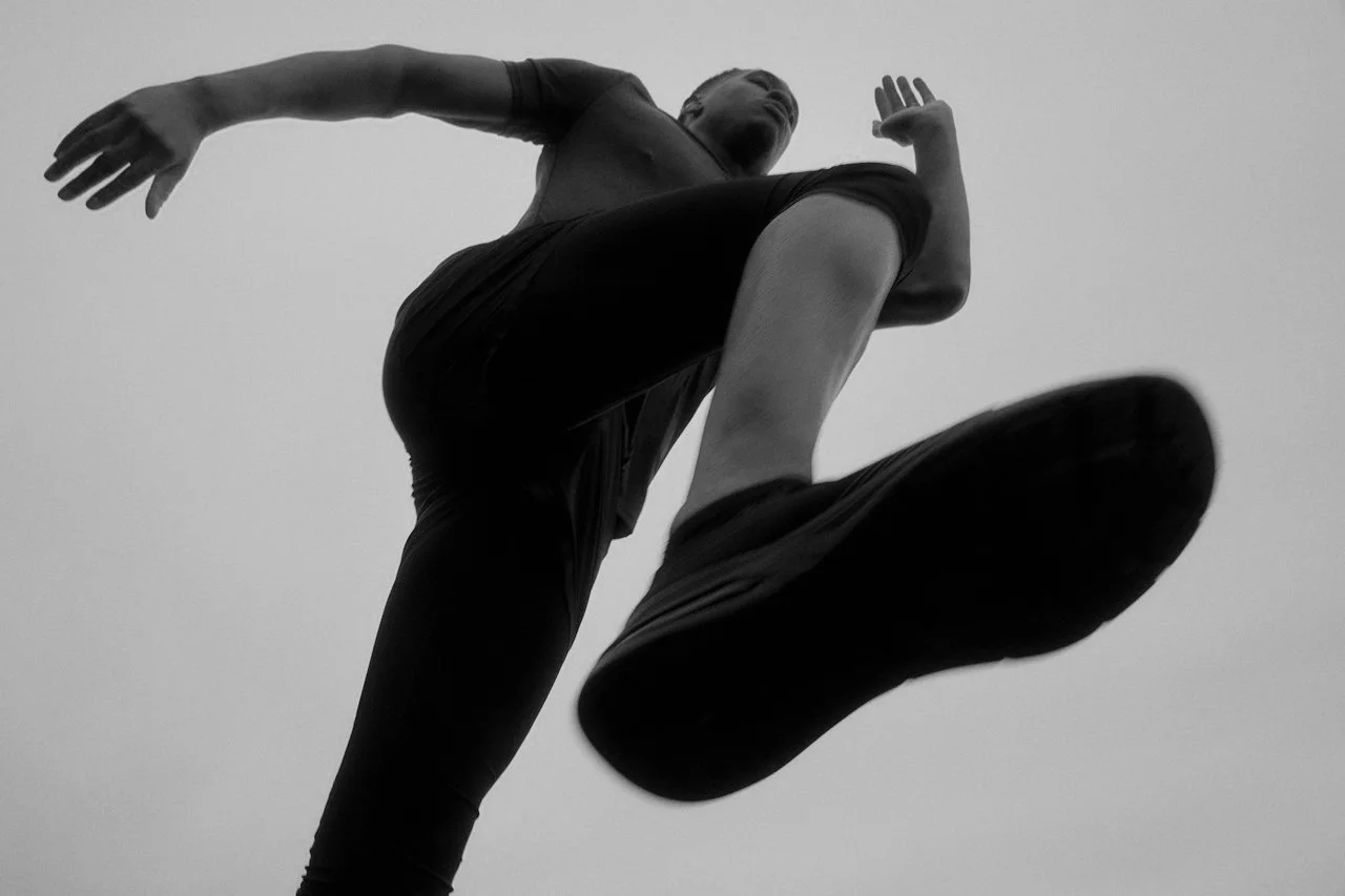 A person in athletic clothing is shown from below in a black and white photo, capturing them mid-air, possibly jumping or kicking against an overcast sky.