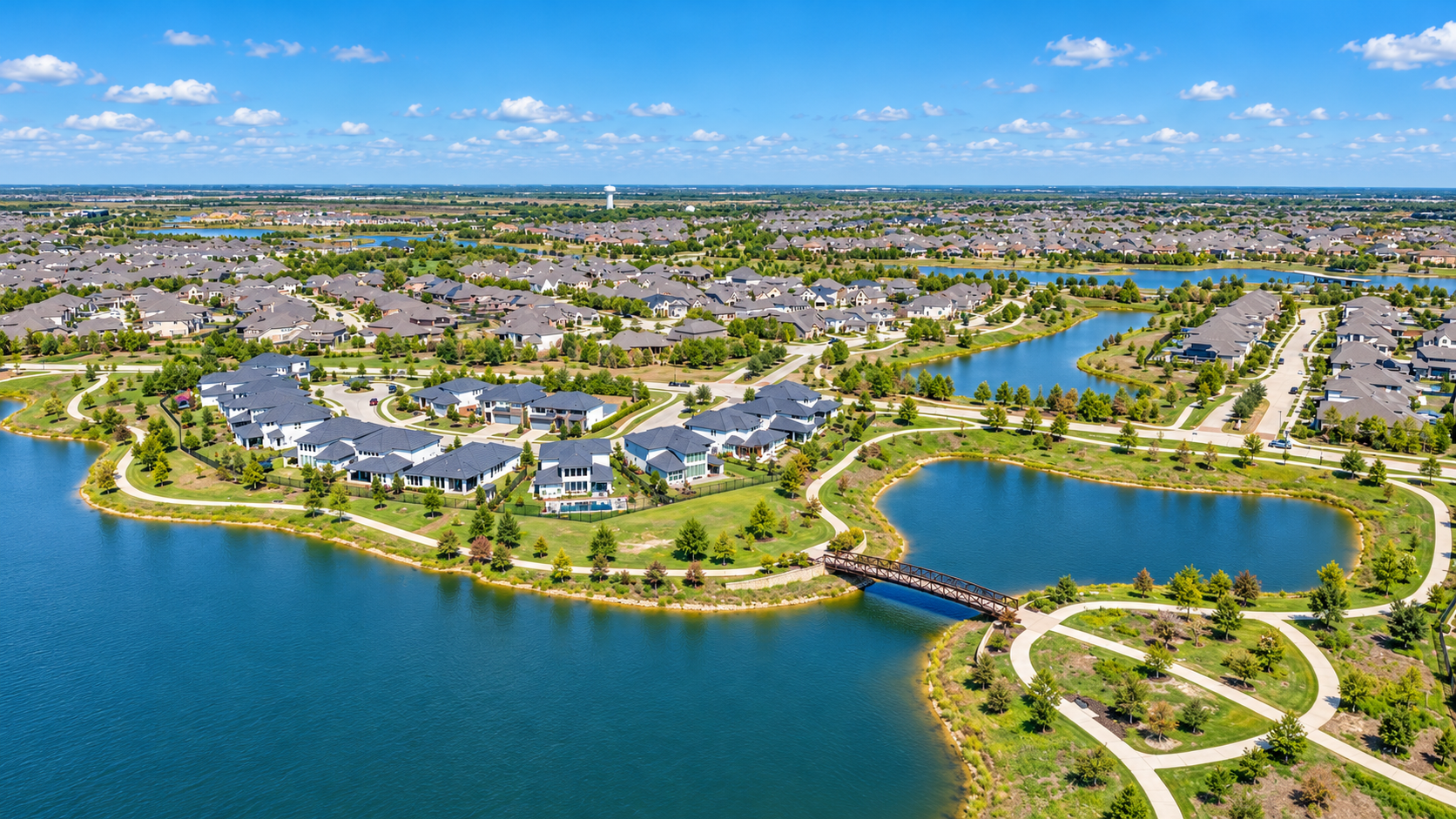 Aerial view of a suburban neighborhood with lakes, houses, green lawns, and trees under a blue sky with clouds.