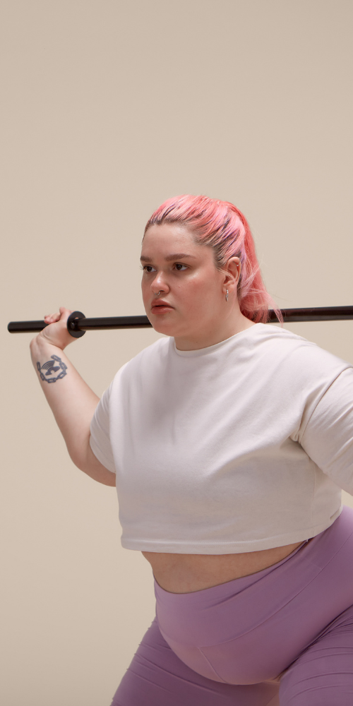 Eine übergewichtige Frau mit pinkfarbenen Haaren hält eine Langhantel hinter dem Kopf, steht vor einer hellen Wand und trägt ein weißes T-Shirt und lilafarbige Leggings.