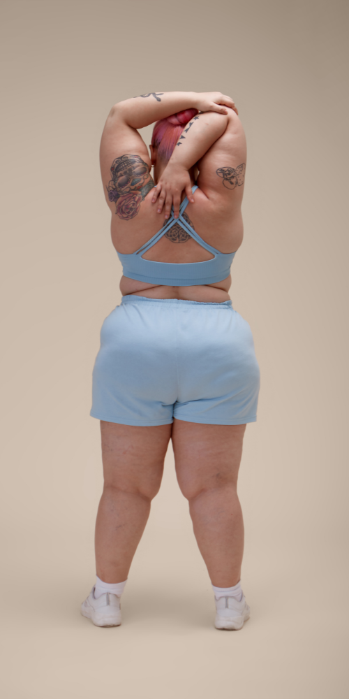 Ein Mensch mit Tattoos auf dem Arm und Rücken, trägt einen blauen Sport-BH und Shorts, steht vor neutralem Hintergrund, die Arme über dem Kopf verschränkt.