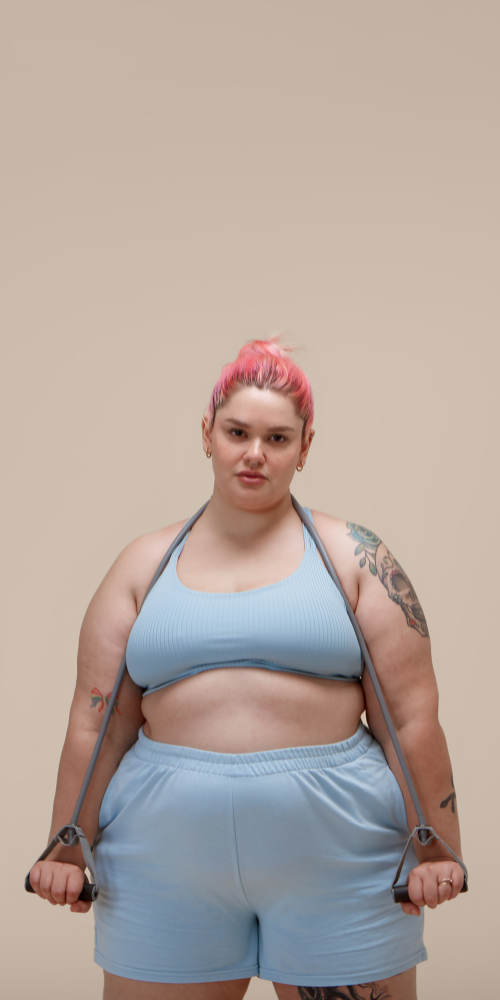 Junge Frau mit pinkem Haarknoten trägt graues Sporttop, Shorts und Springseil, steht vor neutralem Hintergrund.