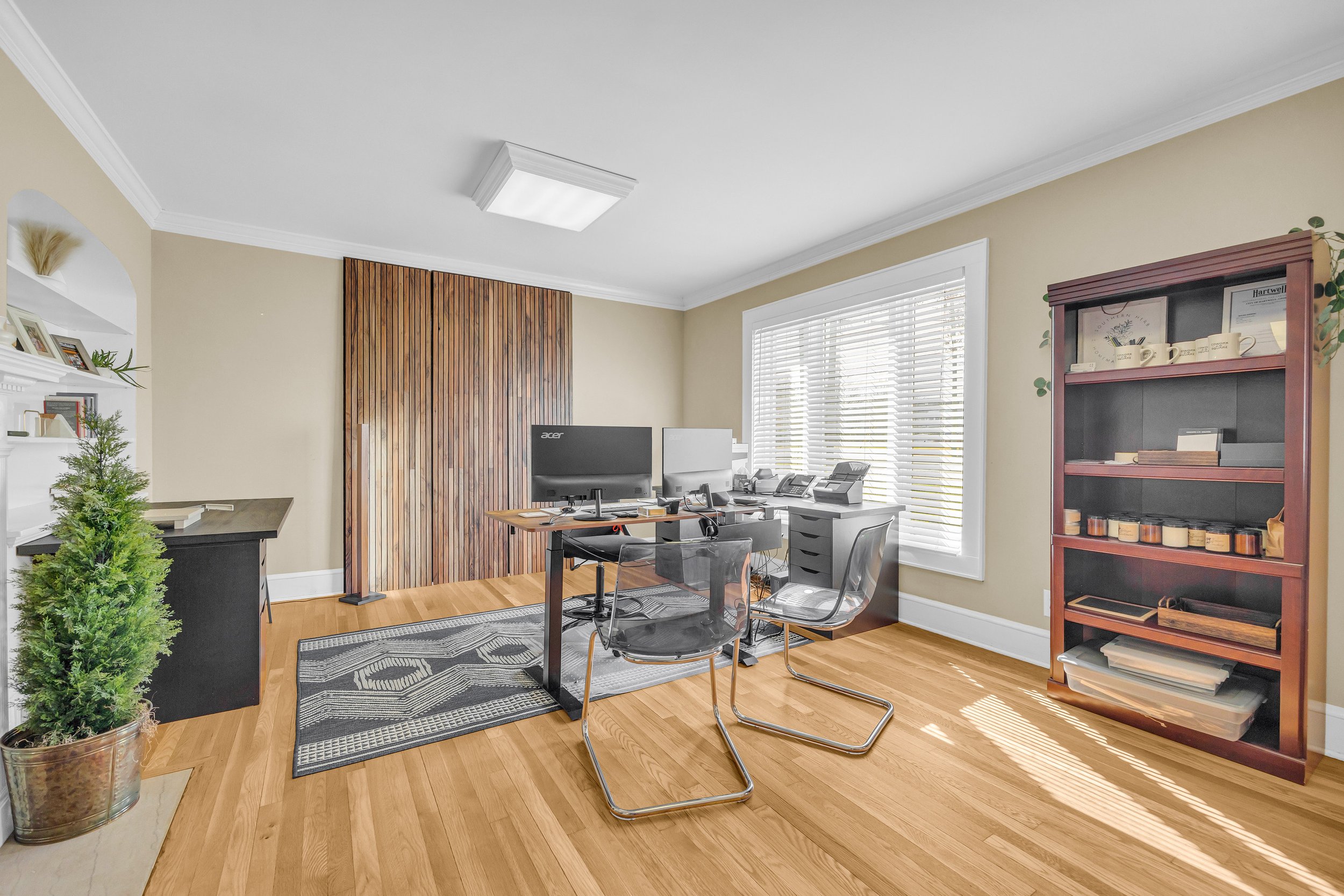 Modern home office with wooden floors, a desk with dual monitors, transparent chairs, a large window with blinds, and a bookshelf.