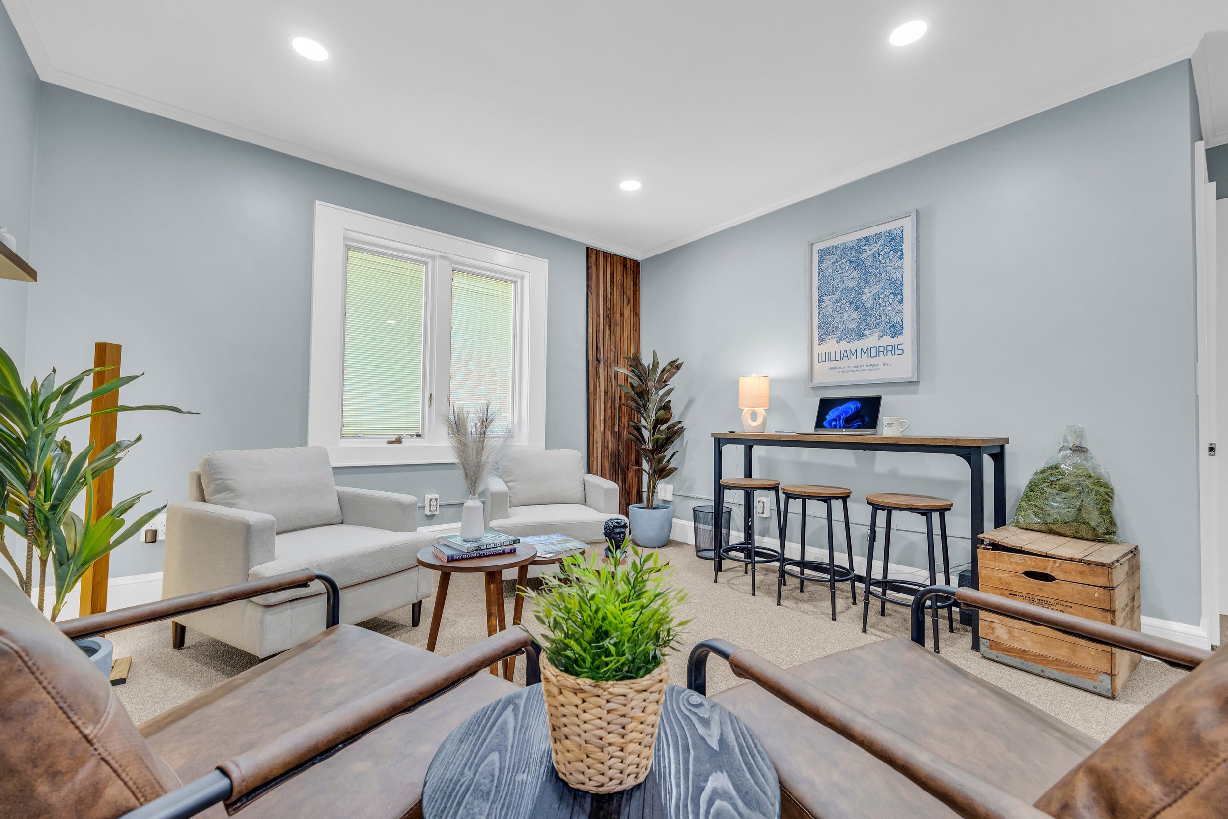 Modern living room with beige armchairs, wooden coffee table, decorative plants, wall art, and a bar-style table with stools against a light blue wall.