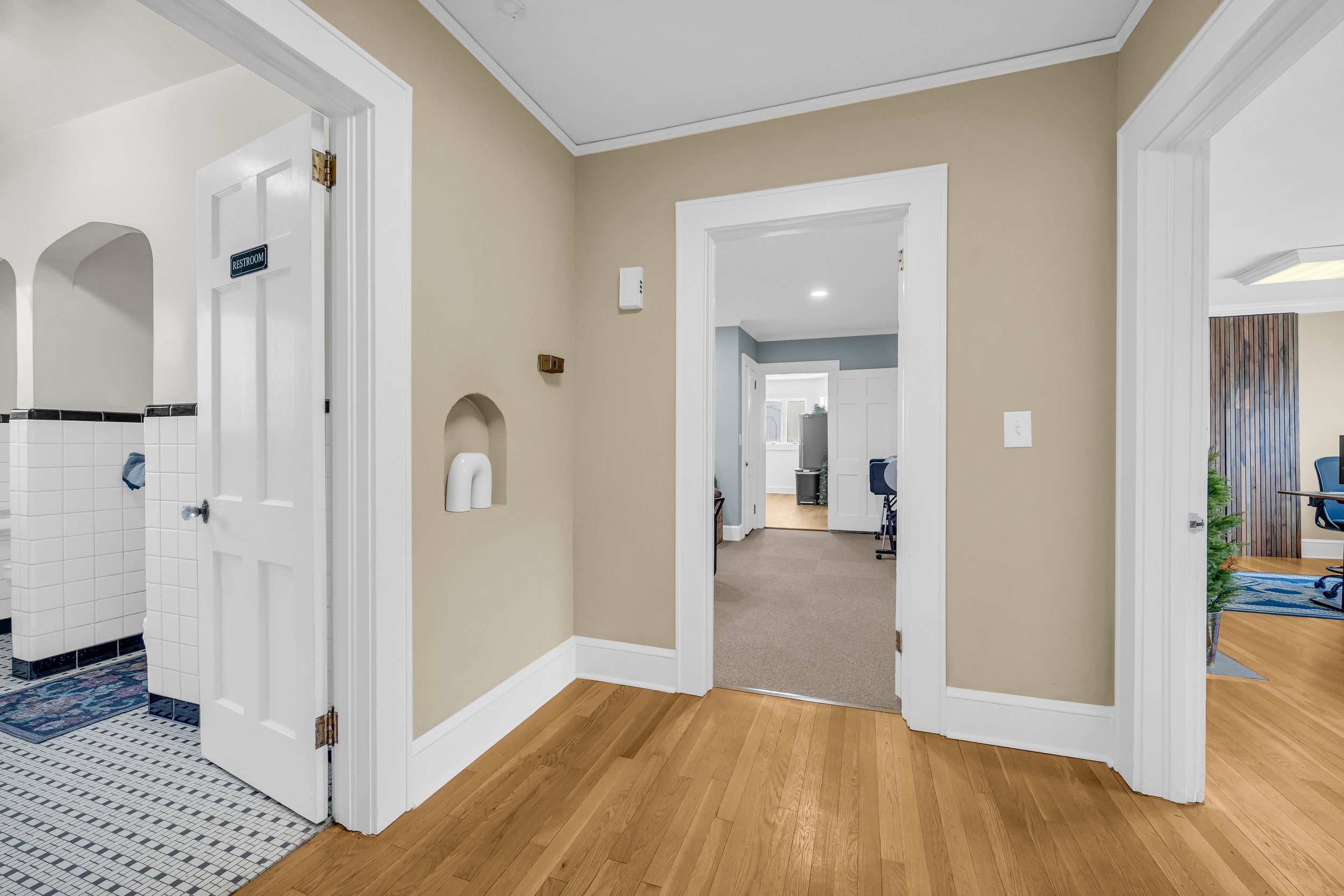 Interior view of a hallway with light-colored walls and wooden flooring. The image shows open doorways leading to a tiled restroom and other rooms with carpeting. The restroom has white tiled walls and a blue and white patterned floor. Office chairs