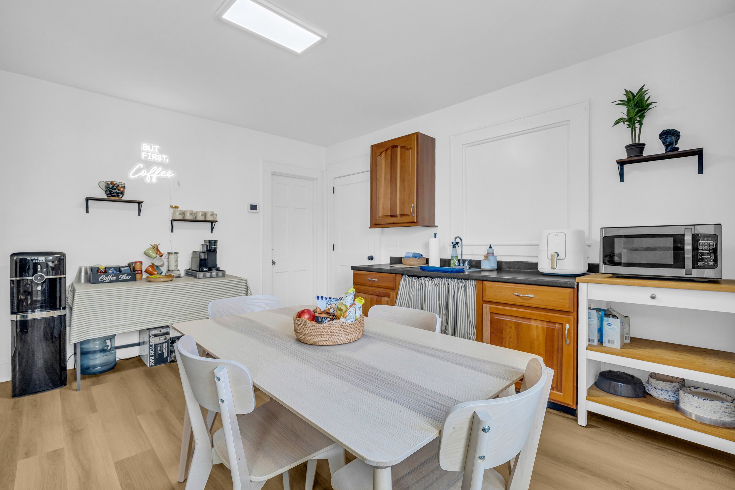 Modern kitchen with a wooden table, chairs, microwave, air fryer, and coffee station with neon sign.