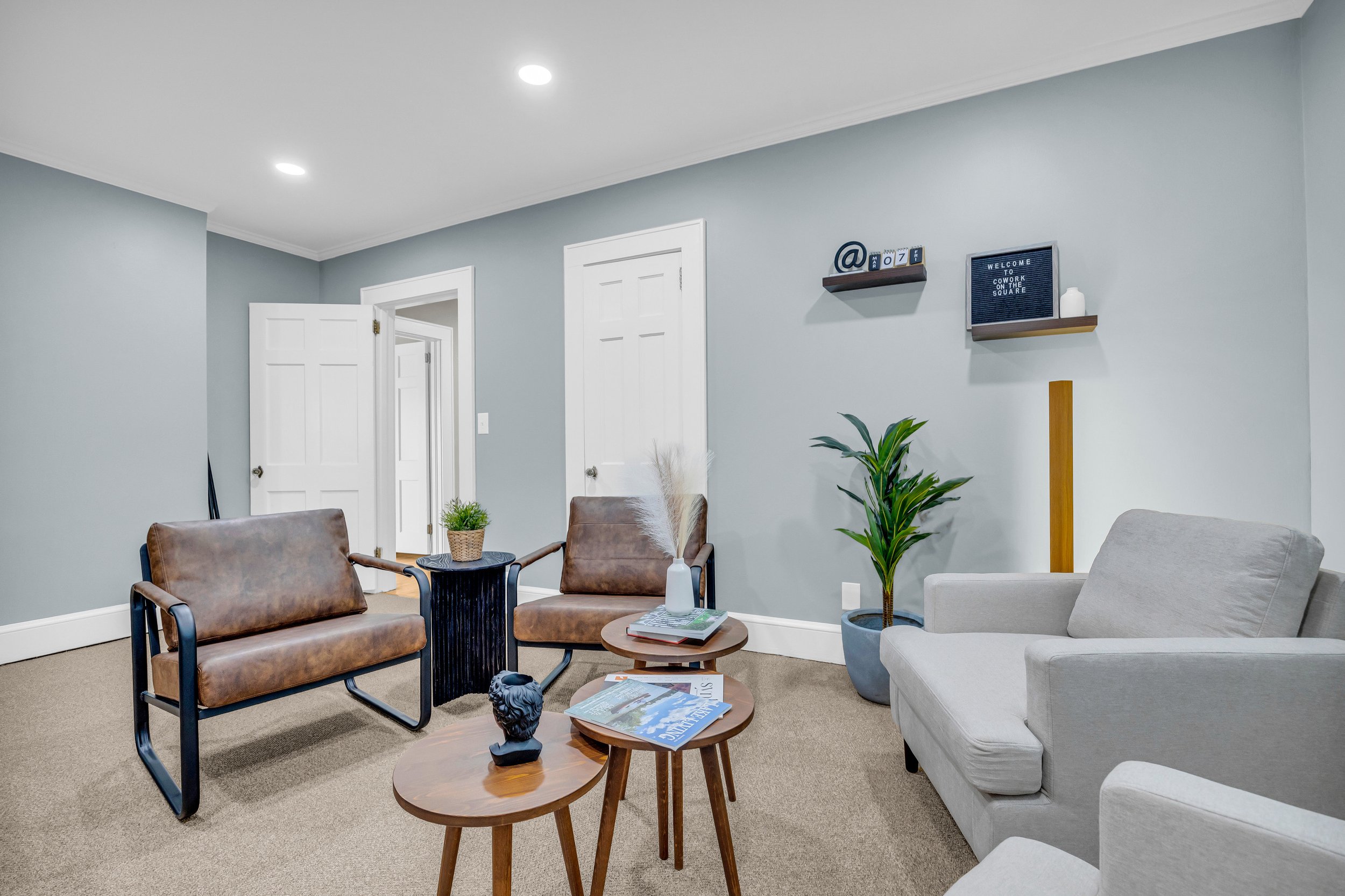 Modern office lounge area with light blue walls, two brown leather chairs, a light gray armchair, nested wooden tables, a plant in a pot, and decorative items on a black side table. Shelves on the wall hold a welcome sign and decorative objects.