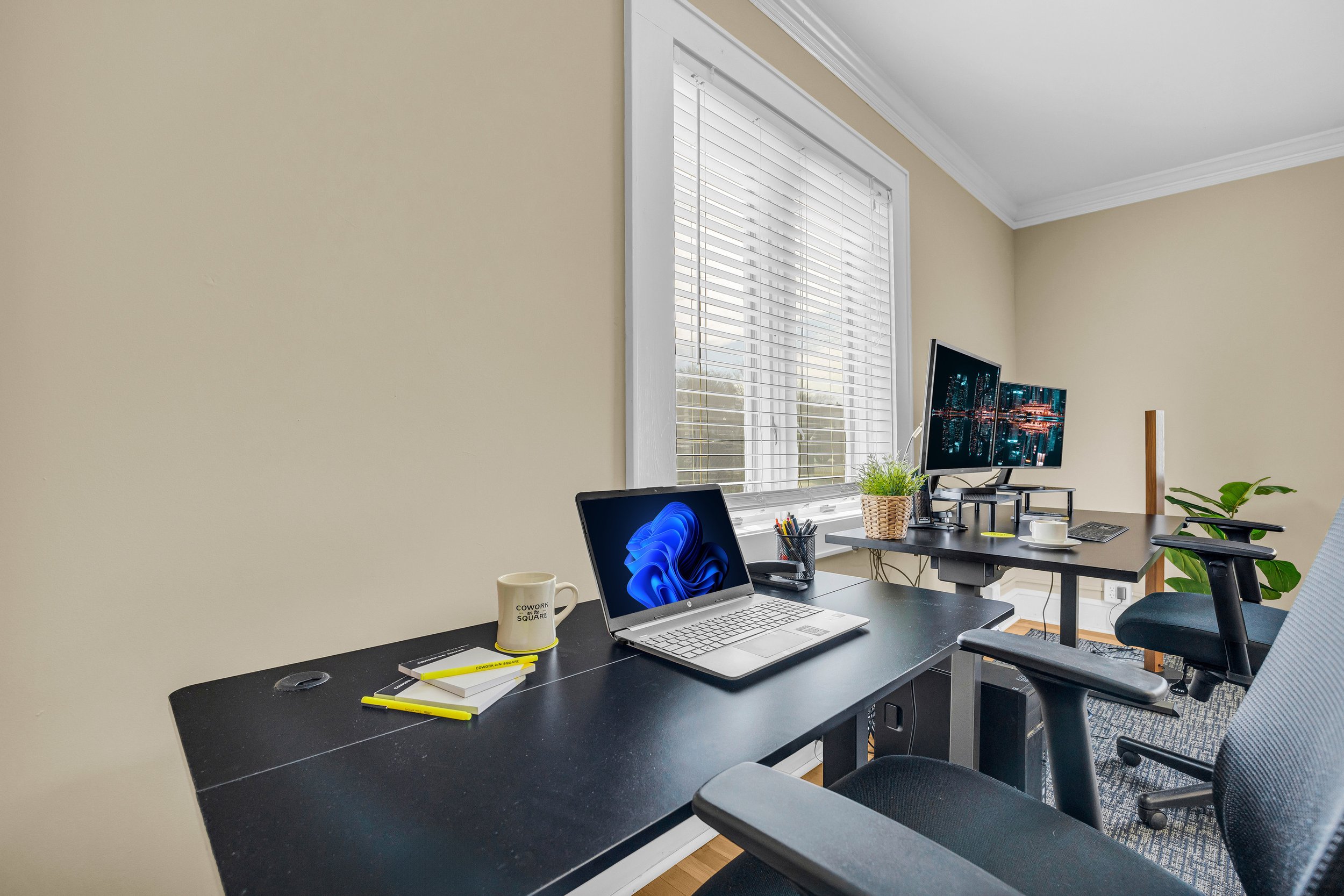 Home office with black desk, laptop, dual monitors, chair, potted plant, and window blinds