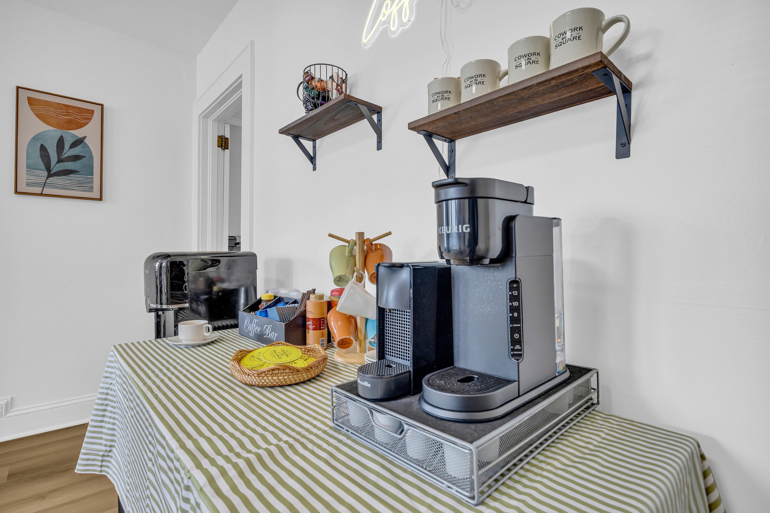 Home coffee station with a Keurig coffee maker, assorted mugs, toaster, and coffee supplies on a striped tablecloth, with shelves and a decorative art piece on the wall.
