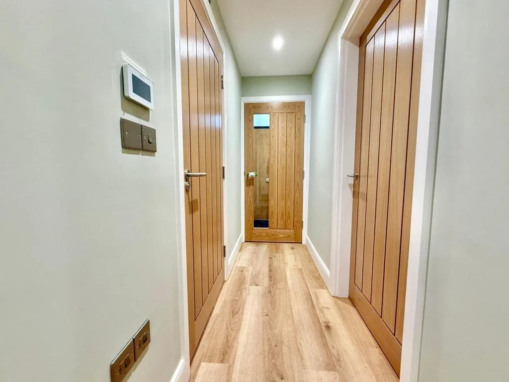 A hallway with light-colored wooden doors and LVT flooring, including a door with a small window at the end, and an underfloor heating control on the left wall.