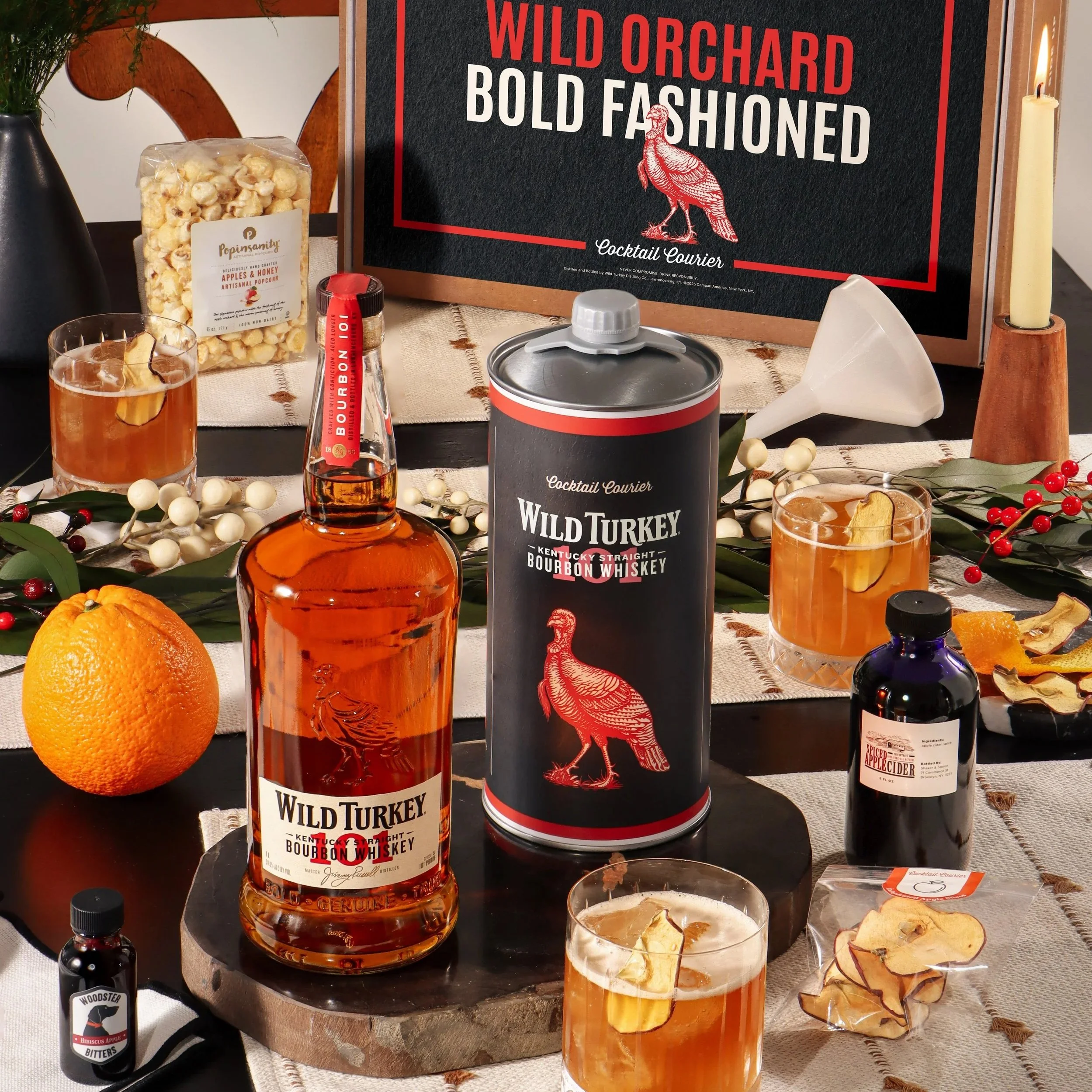 A table display with a large sign reading 'Wild Orchard Bold Fashed.' There are bottles of whiskey, a glass of whiskey with a lemon slice, popcorn, a candle, and decorative berries and leaves on the table.