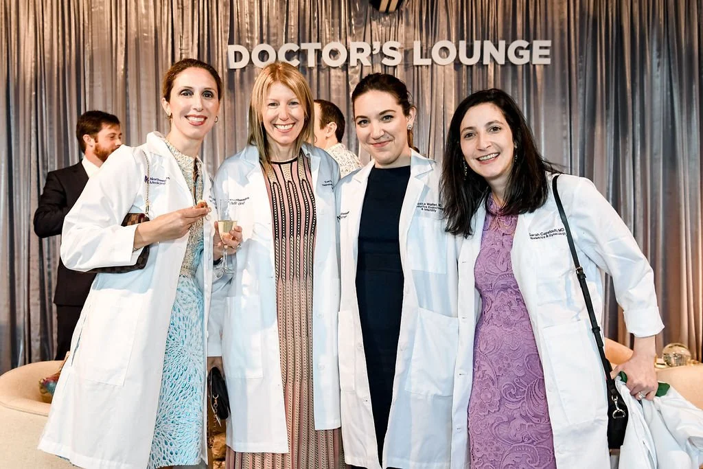 Four women in white medical coats smiling and standing together in a room with a dark curtain backdrop that says 'DOCTOR'S LOUNGE', while two men are in the background at a medical event or conference.