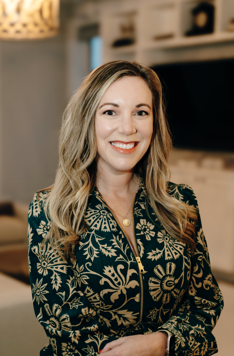 A smiling woman with shoulder-length blonde hair, wearing an off-the-shoulder floral dress, standing outdoors in a park during sunset with trees and grass in the background.