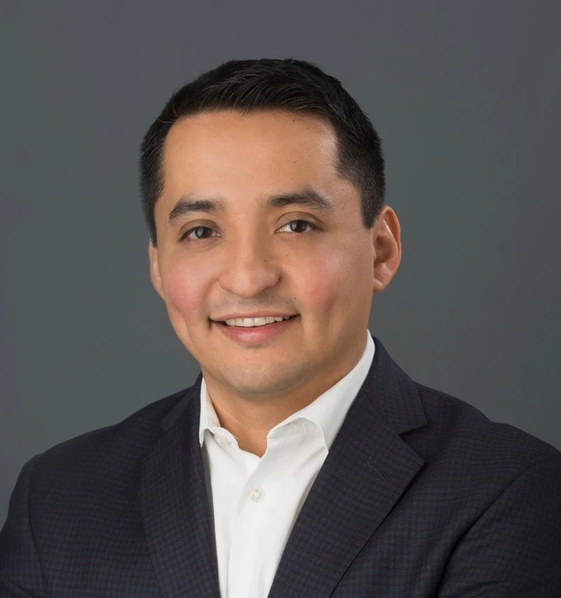 A professional headshot of a man in a black suit and white shirt, smiling with dark hair, set against a gray background.