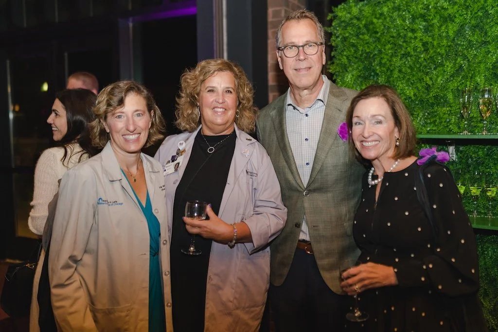 Four people posing together at a social event, two women on the left in beige and light purple coats, one man in a gray checkered blazer, and one woman on the right in a black dress with pearl necklace, all smiling and holding glasses.
