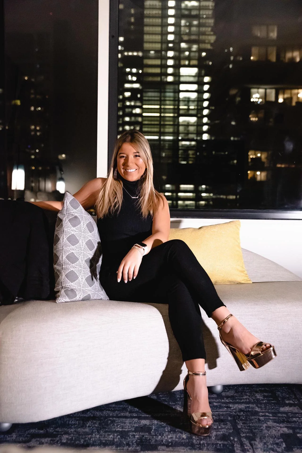 A smiling woman sitting on a beige sofa in a modern apartment at night, with tall city buildings visible through the large window behind her. She is wearing a sleeveless black top, black pants, and gold high-heeled shoes, with her hair down.