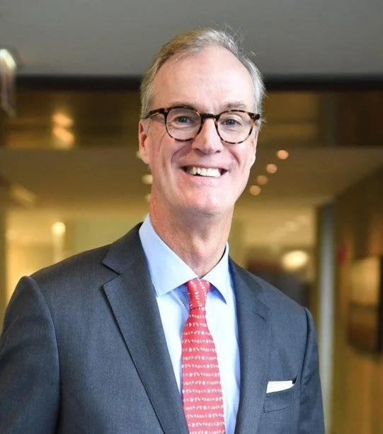 A smiling man in a gray suit, light blue shirt, and patterned red tie, wearing glasses, standing in an indoor setting with blurred background lighting.