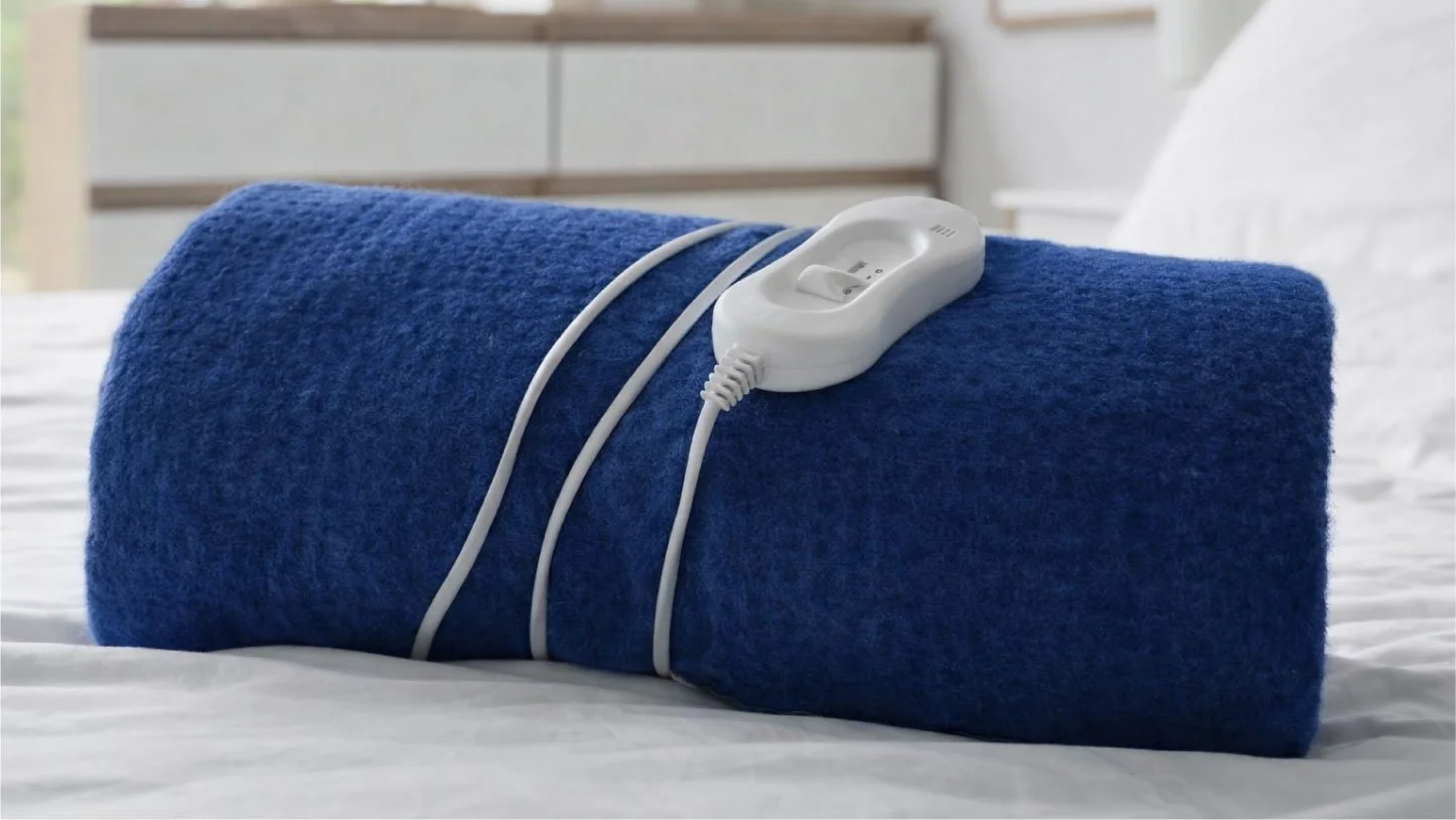 Rolled blue heating pad with attached controller resting on a bed in a calm bedroom setting.
