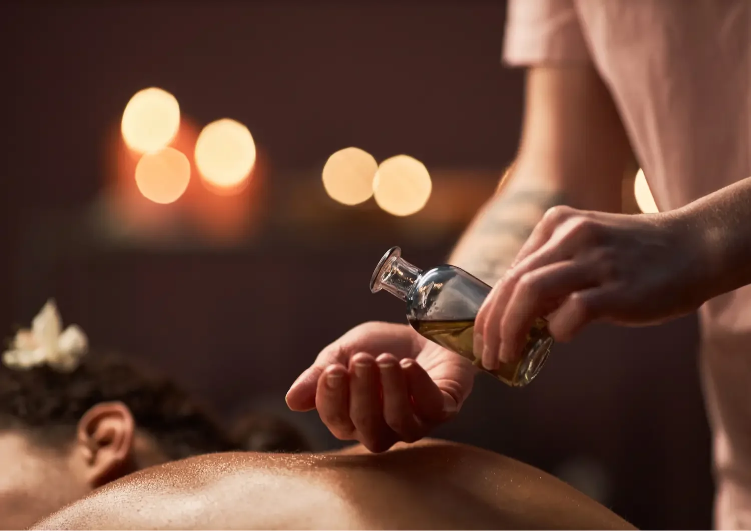 Massage therapist pouring oil on a person's back in a dimly lit setting with blurred warm lights in the background.