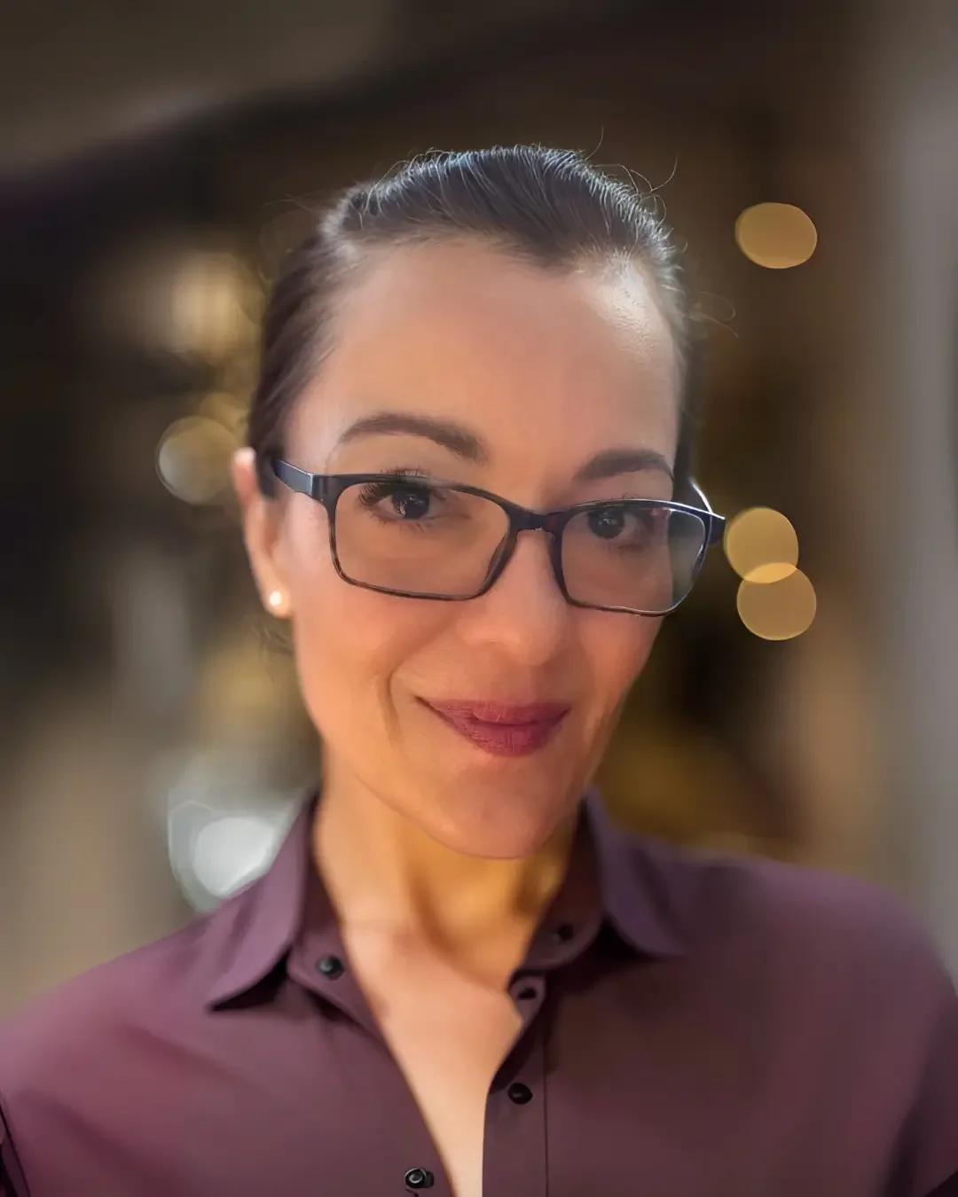 A woman with dark hair, wearing glasses and a maroon blouse, smiling at the camera with a blurred background and circular bokeh lights.