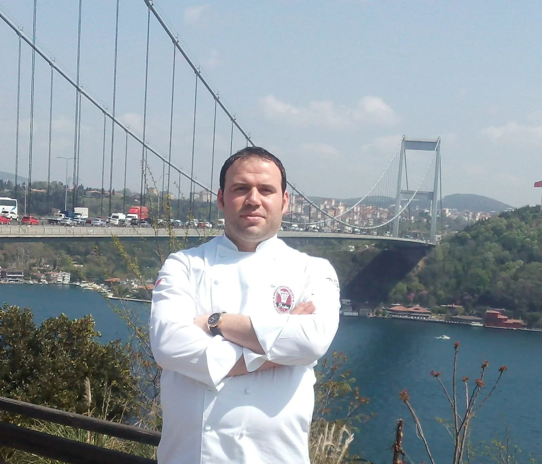 Önde, beyaz bir şef kıyafeti giymiş erkek, kolları çapraz, duruyor. Arka planda Boğaziçi Köprüsü ve bir gölet veya boğaz var.