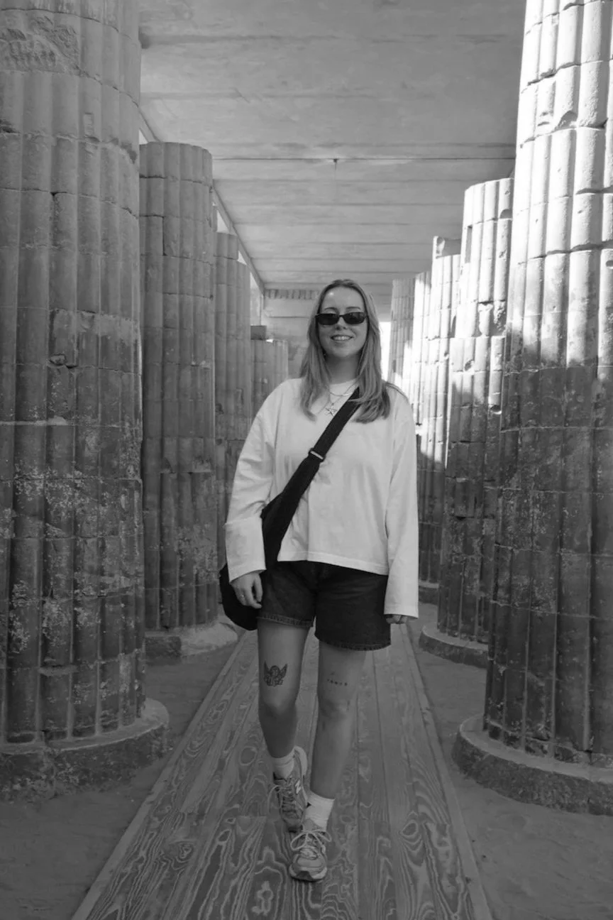 A young woman smiling and wearing sunglasses, a white top, and shorts, standing on a wooden pathway between ancient stone columns in a historical site.