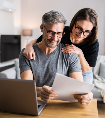 A middle-aged man and a woman with glasses looking at a paper together, with the woman hugging the man from behind in a cozy living room.