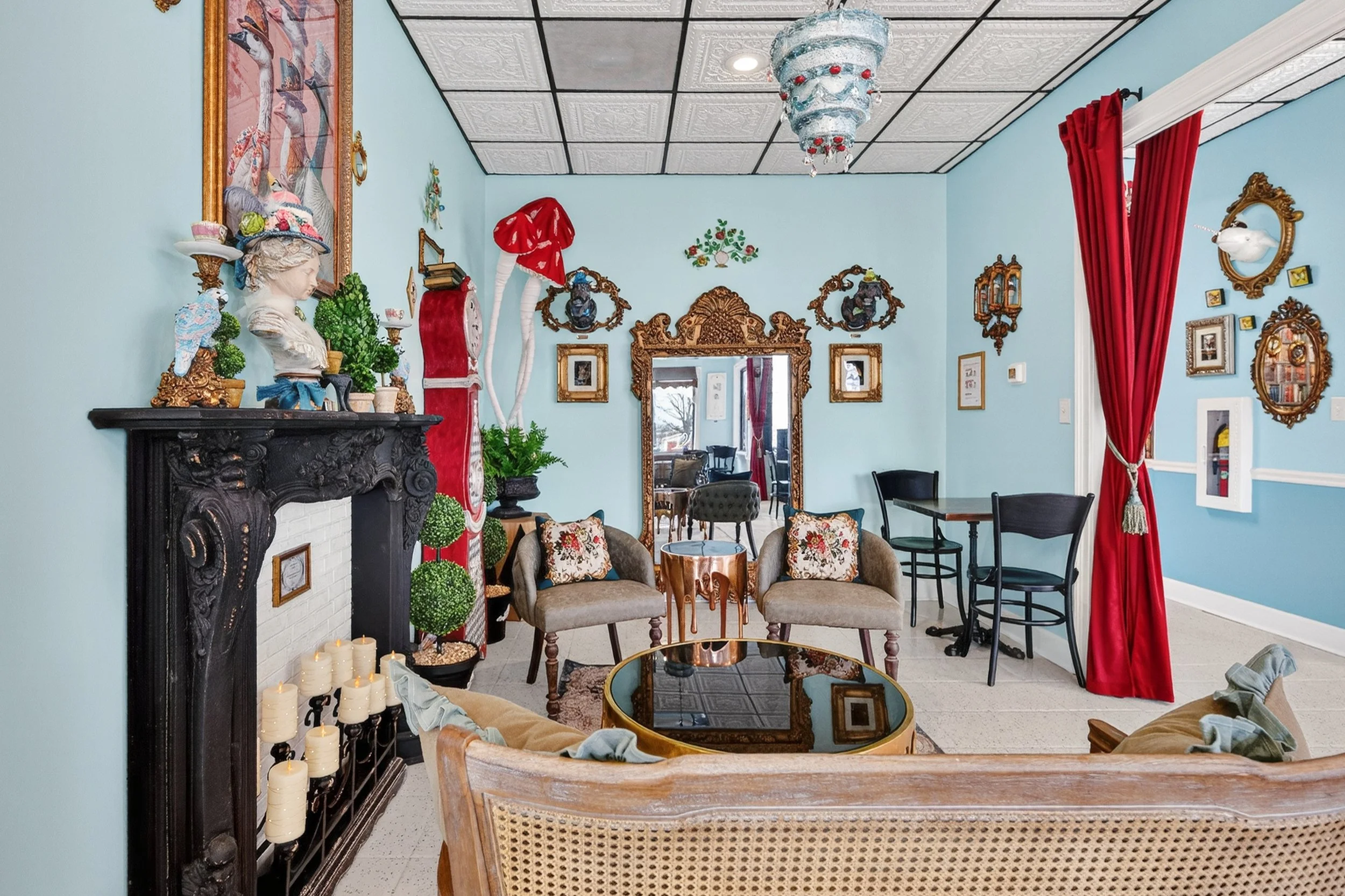 A vintage-style room decorated with eclectic art, sculptures, and furniture. Features a black ornate fireplace mantel, a large mirror with a carved frame, two armchairs with floral pillows, a round glass-top table, and red curtains. The walls are lig