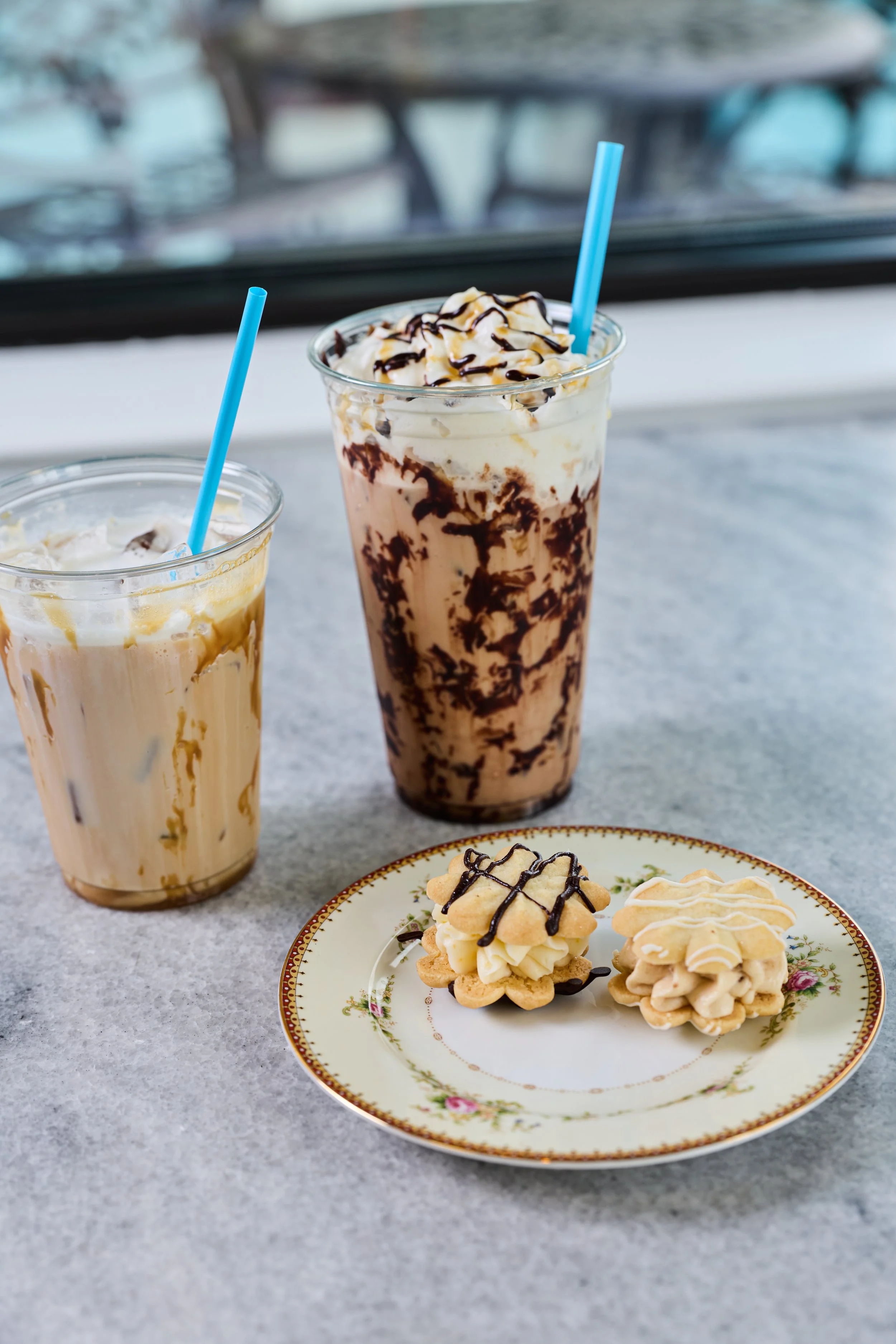 Two large iced coffee drinks with whipped cream and chocolate drizzle. In front, a small plate with two butter cookies.