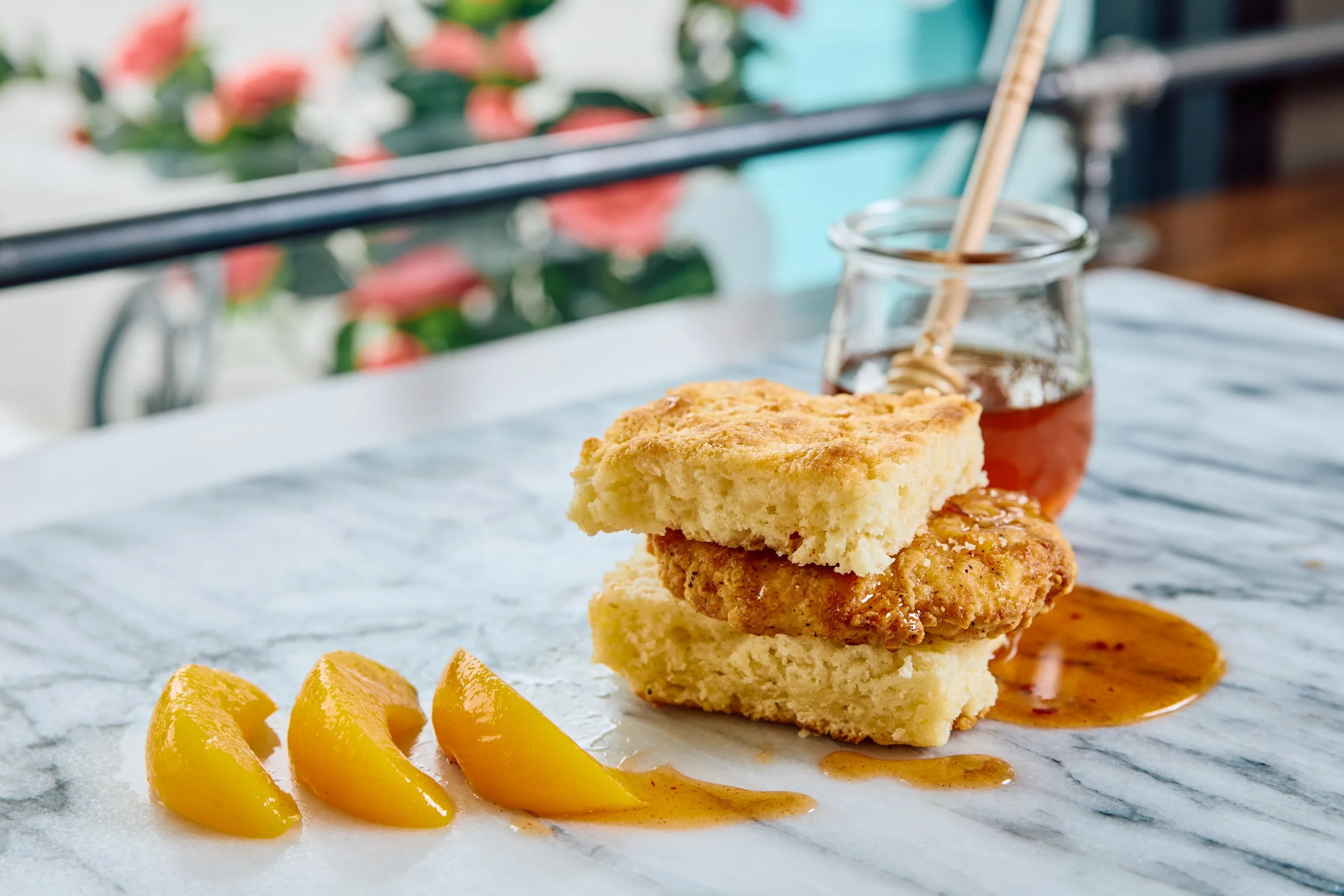 A breaded chicken breast between a biscuit, with slices of peach and a jar of honey.
