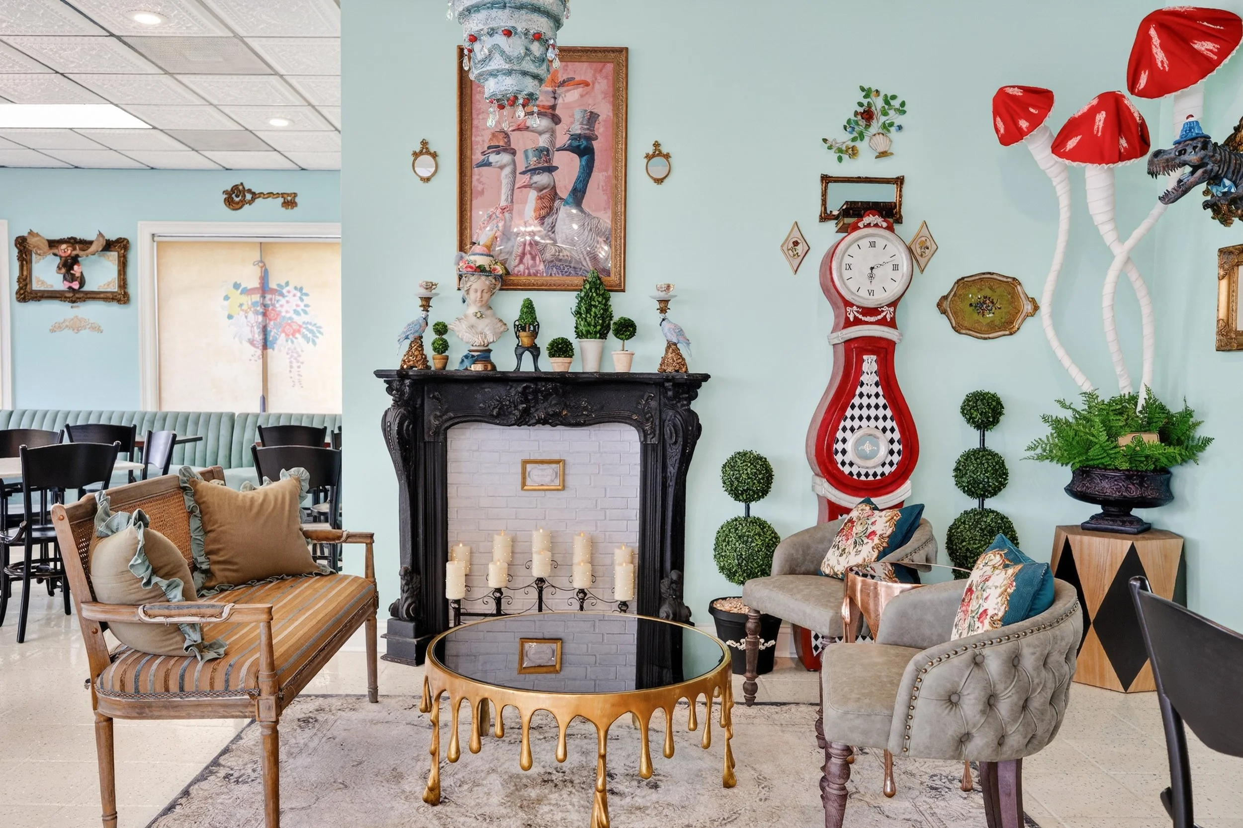 A cozy indoor seating area with vintage and eclectic decor. It features a black fireplace with candles, a sofa, a tufted armchair with floral pillows, a round black-and-gold coffee table, and mushroom and dinosaur sculptures on the wall. Decorative p