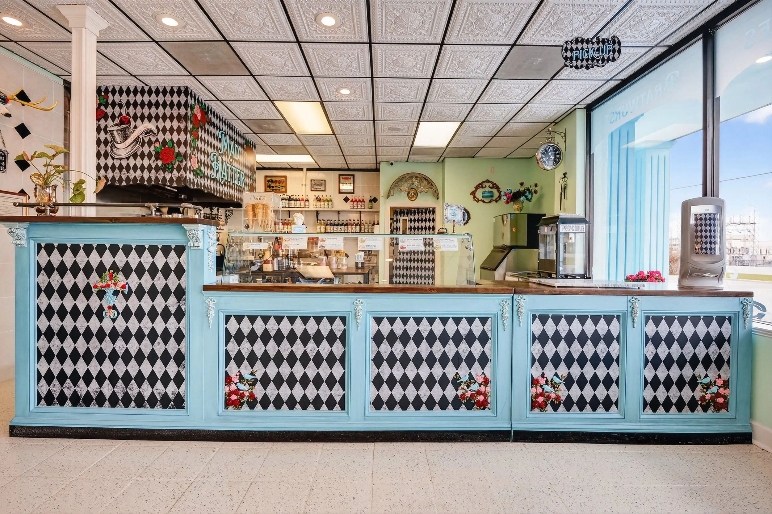 Inside a vintage-style ice cream or dessert shop with a blue and black harlequin pattern counter, pink floral accents, and decorative wall art. The shop has a glass display case, various bottles, and a colorful sign that says 'Mad Platter.' There is 