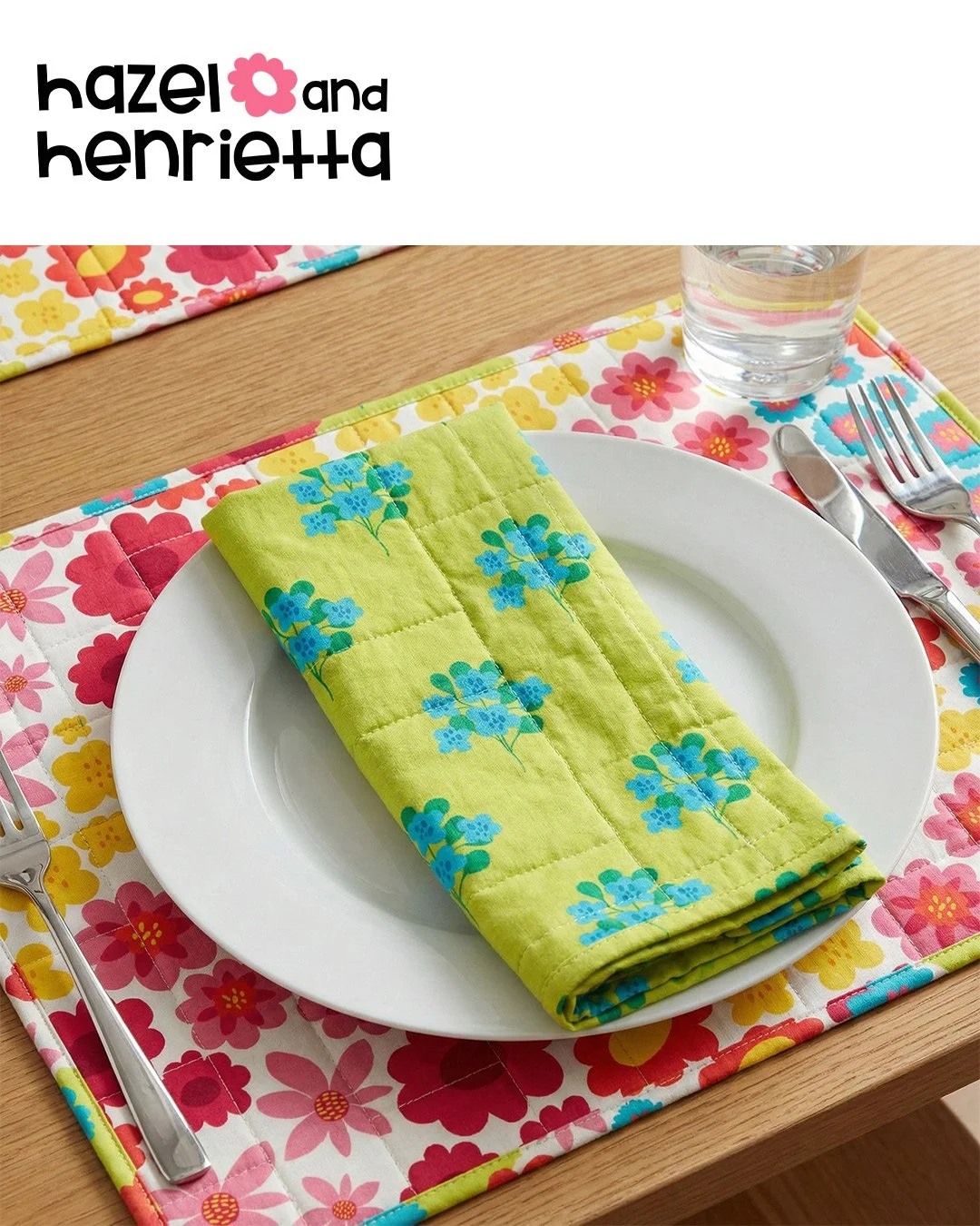 Table setting with a white plate, green floral napkin, silverware, a cup of water, and floral placemats with pink, red, yellow, and blue flowers on a wooden table.