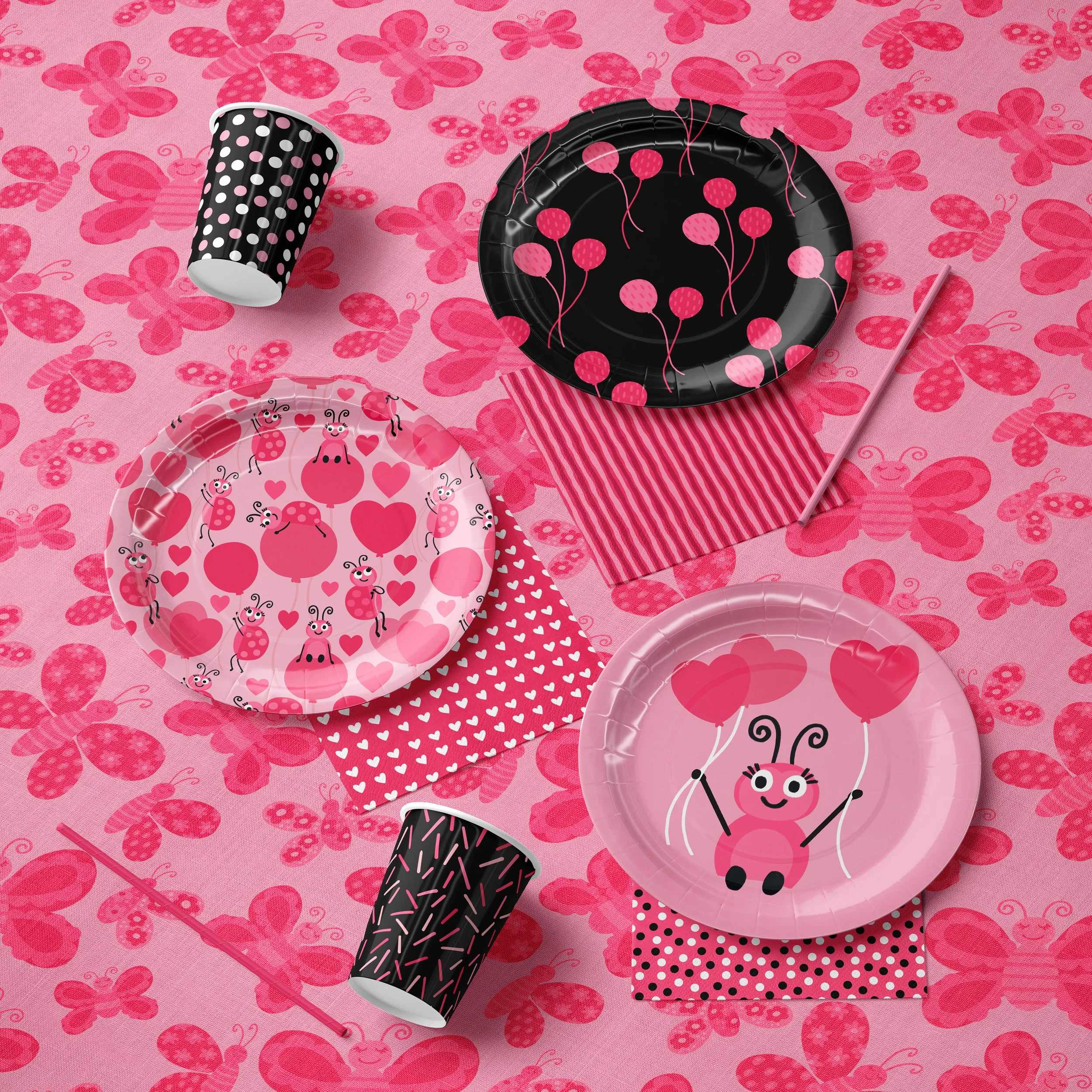 Pink and black butterfly-themed party table setting with paper plates, cups, striped and dotted napkins, and pink and black balloons on a pink butterfly-patterned tablecloth.