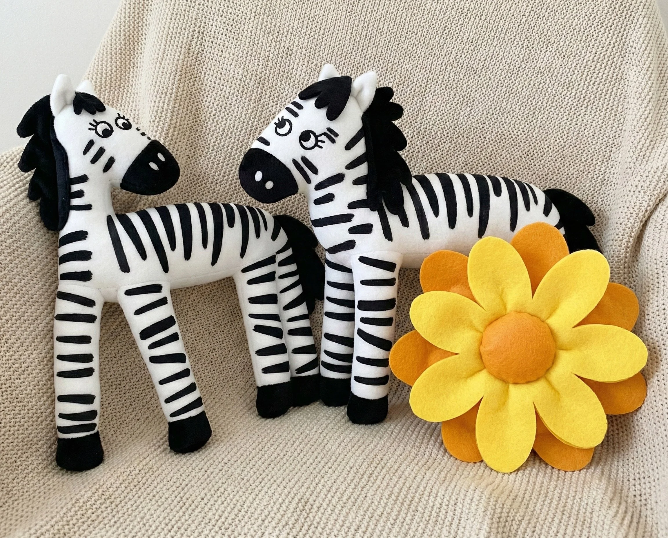 Two plush zebra toys with black and white stripes and a yellow felt flower with orange accents on a beige textured surface.