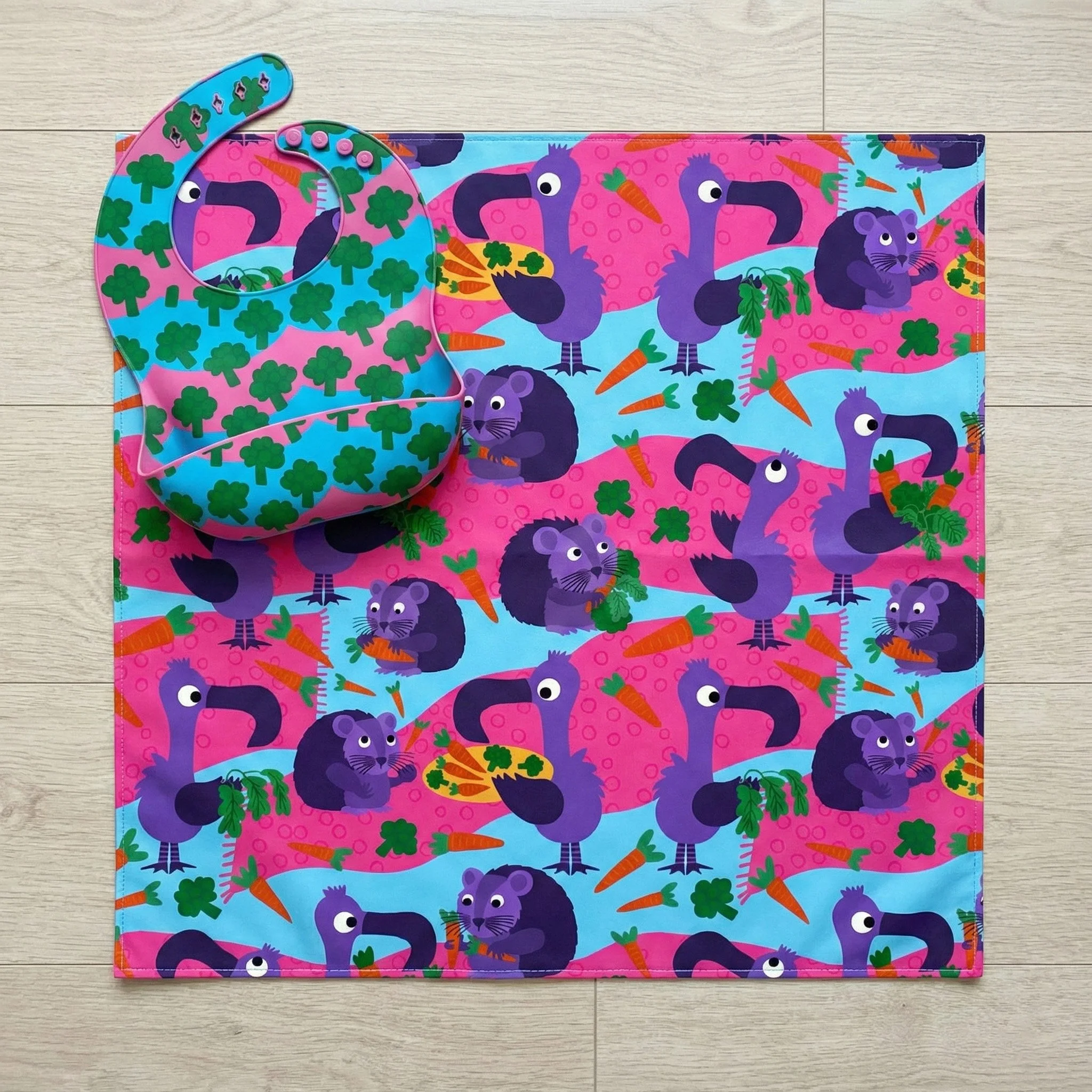 Whimsical bib and splash mat set featuring playful vegetable-loving animals from the Eat Your Veggies collection. This vibrant design showcases cheerful birds and curious critters surrounded by carrots, broccoli, and garden greens in bold, kid-friend