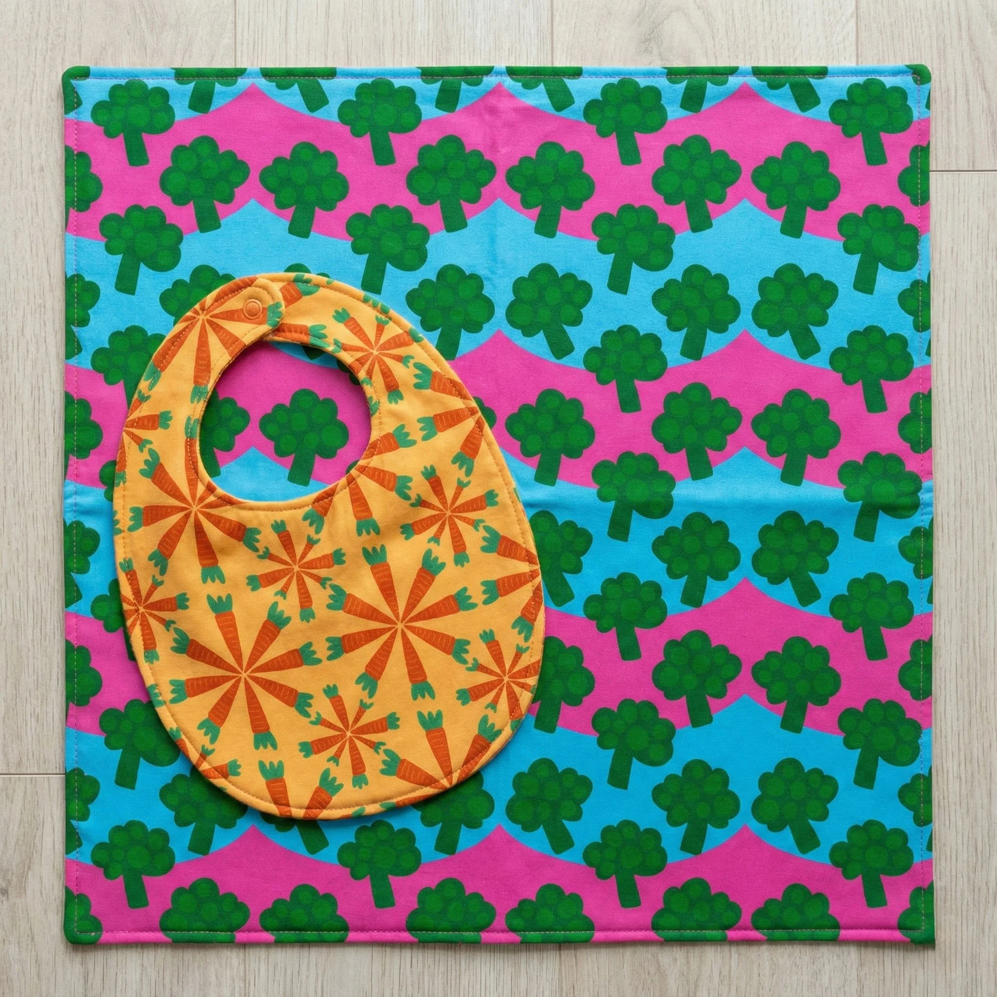 Playful bib and splash mat set featuring bold broccoli and carrot patterns from the Eat Your Veggies collection. Designed with bright colors and whimsical vegetable motifs, this cheerful baby and toddler mealtime set brings fun, creativity, and perso